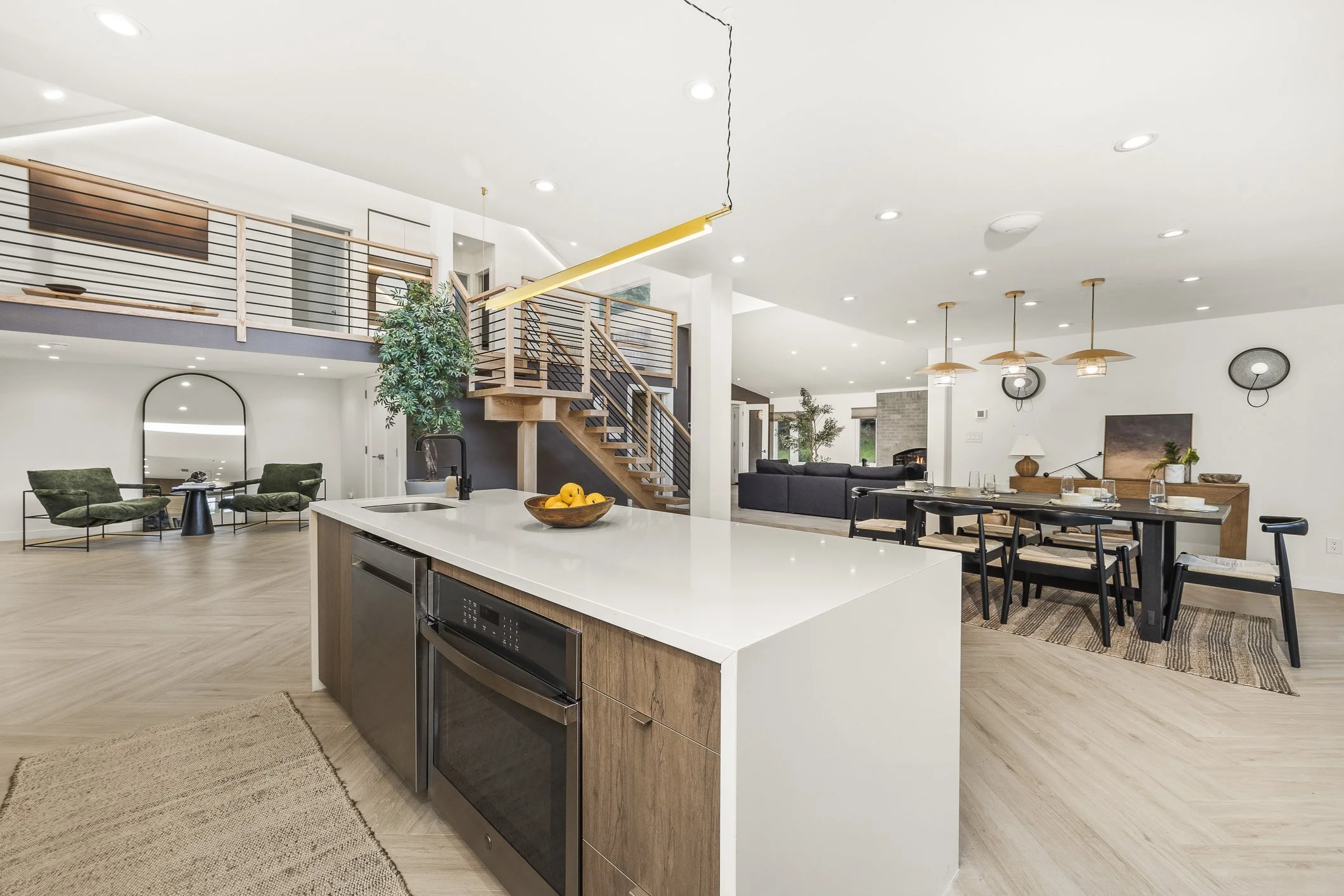 Open-concept living space with kitchen, dining, and living areas, featuring modern furniture, a staircase with a wooden railing, and contemporary lighting fixtures.