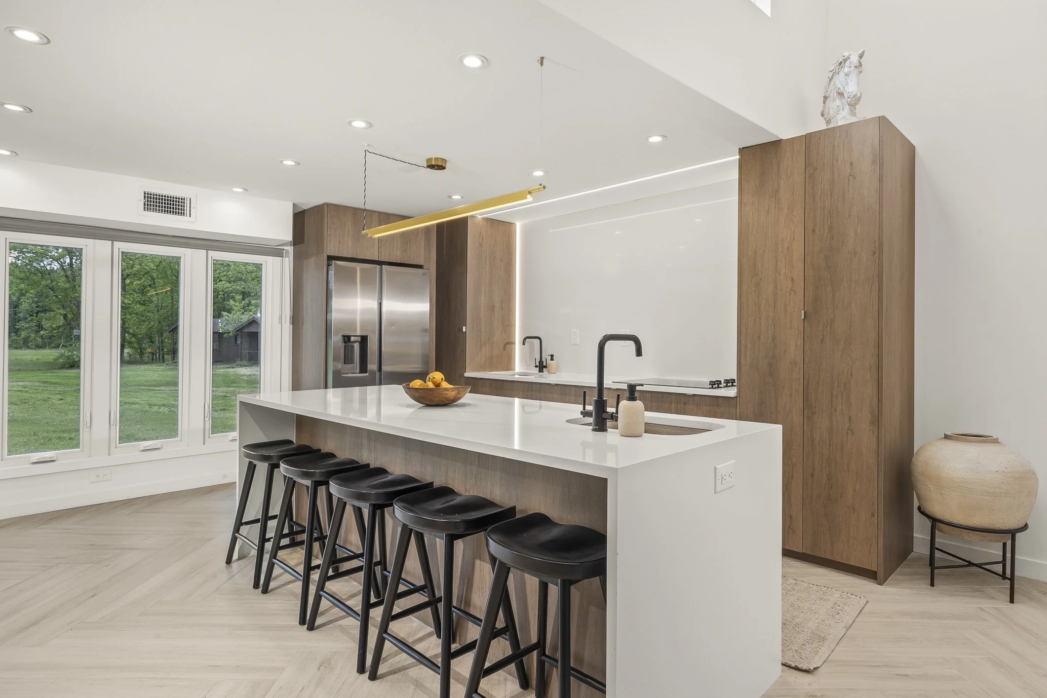 Modern kitchen with white island, black barstools, wooden cabinets, stainless steel refrigerator, and large window with outdoor greenery.