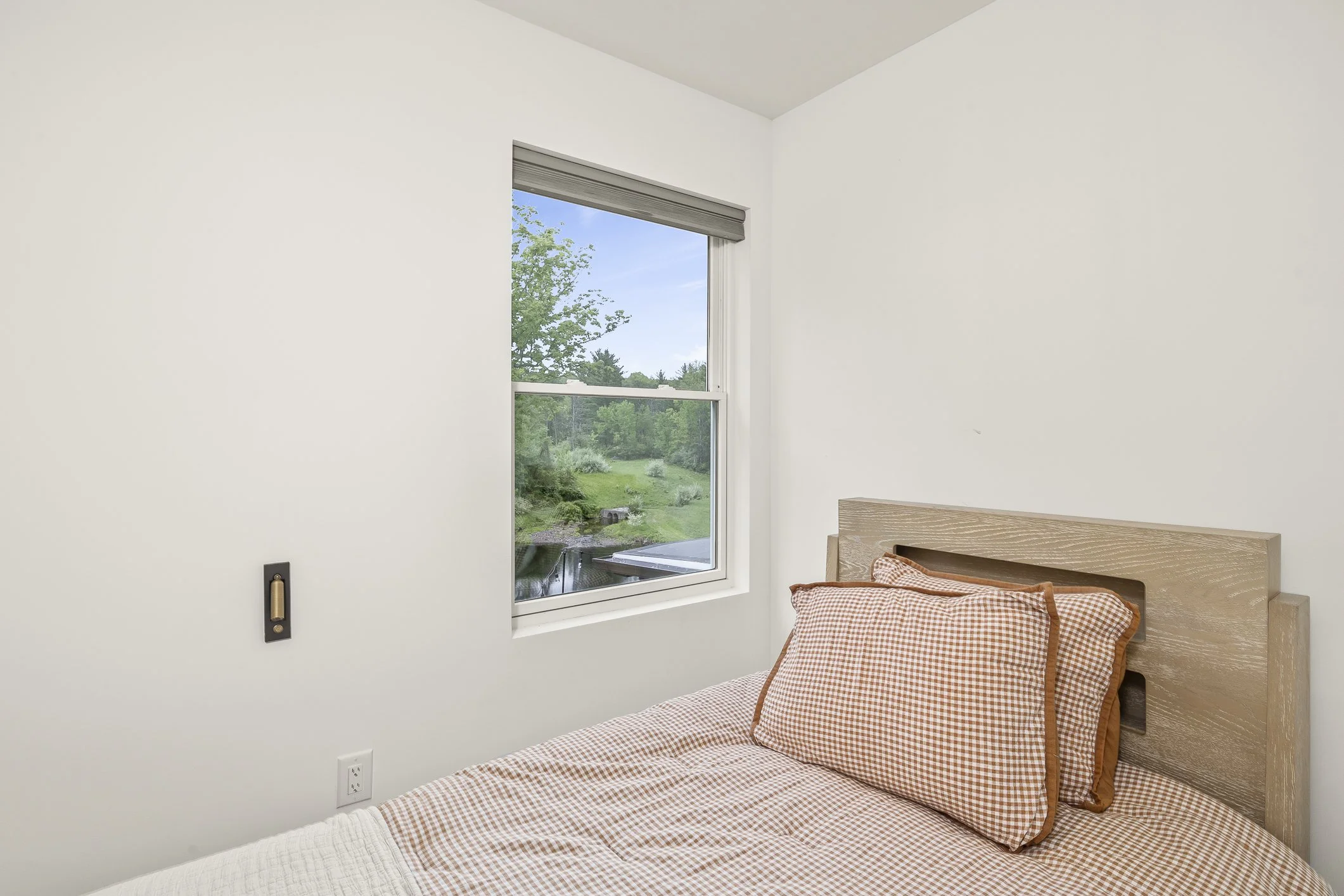 Simple bedroom with a bed covered in orange and white checkered bedding, a wooden headboard, and a window overlooking a lush green outdoor area.