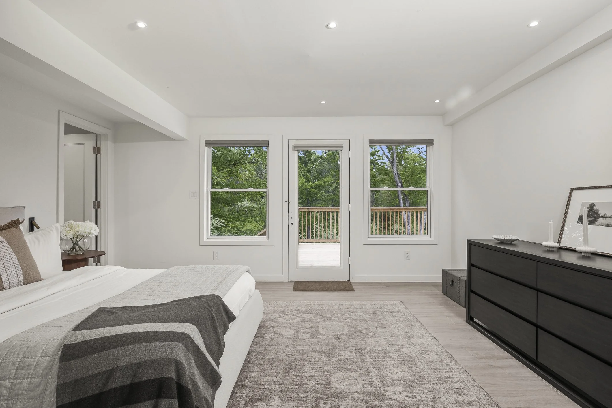 Bedroom with white walls, large window and door leading to balcony with trees outside, bed with gray and white bedding, black dresser with decorative items, neutral-colored rug.