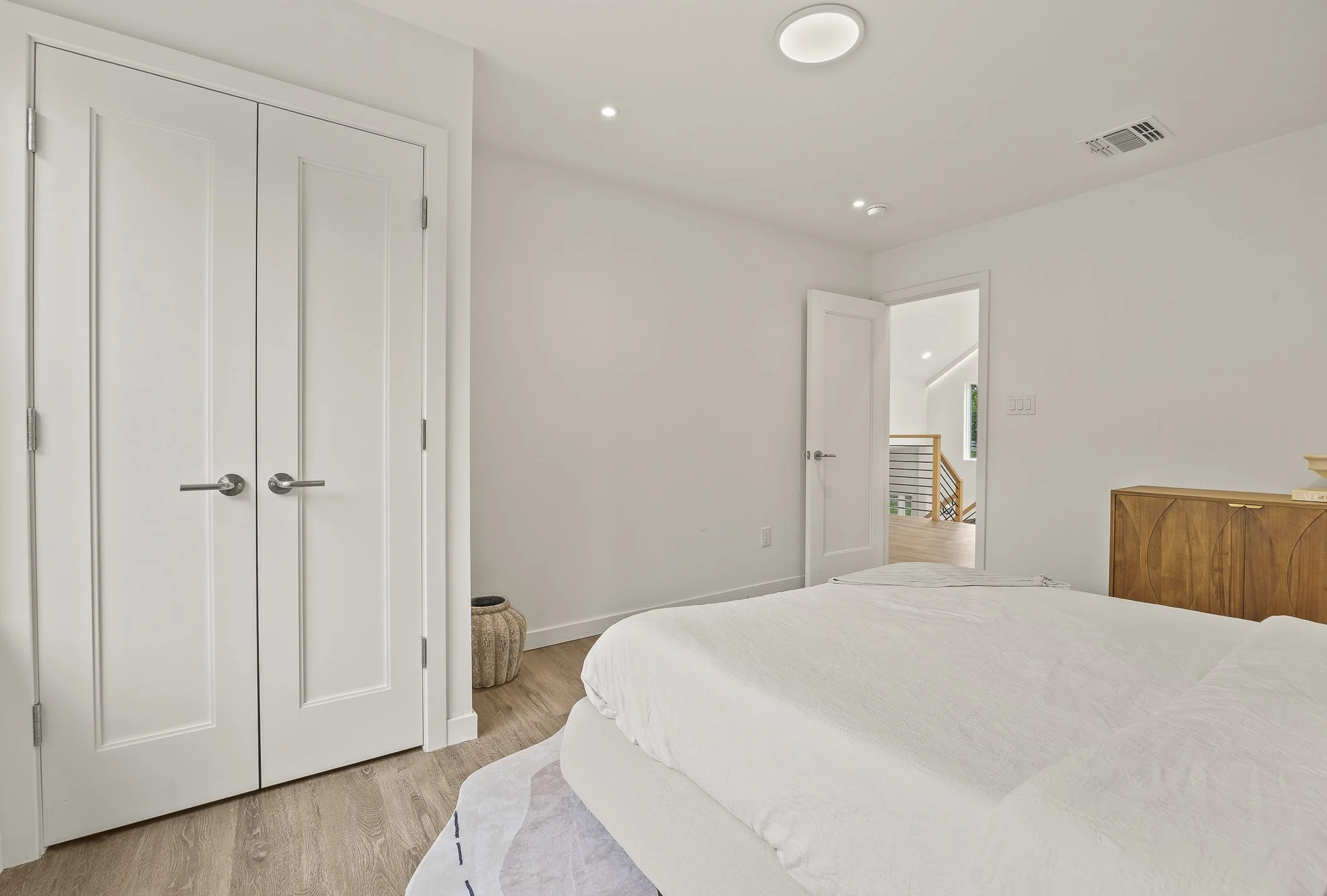 Bedroom with white walls, closed closet doors, a bed with white bedding, a wooden headboard, and an open door leading to a hallway with a staircase and window.