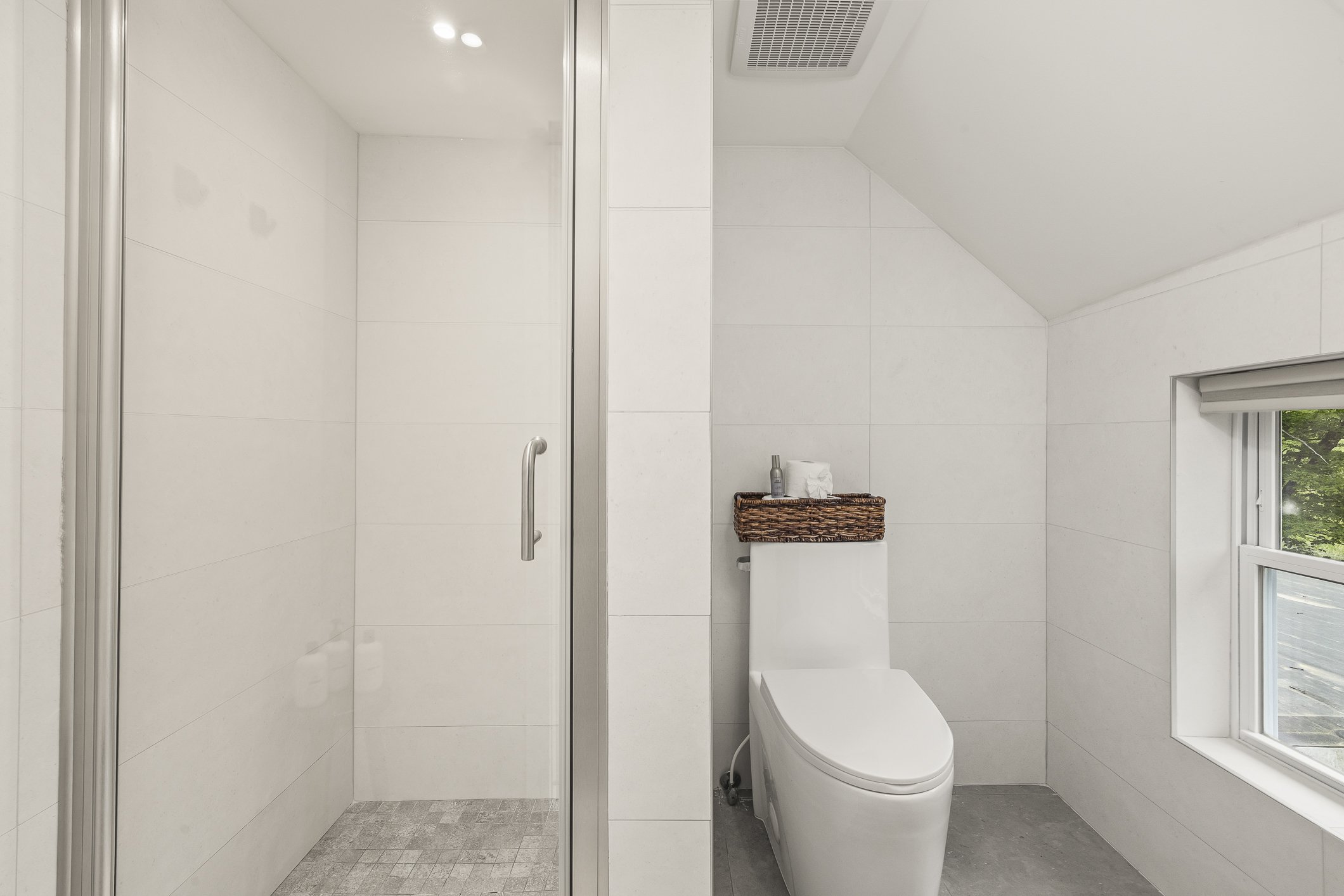 A modern bathroom with a walk-in shower on the left and a toilet with a wicker basket on top on the right, next to a window showing greenery outside.