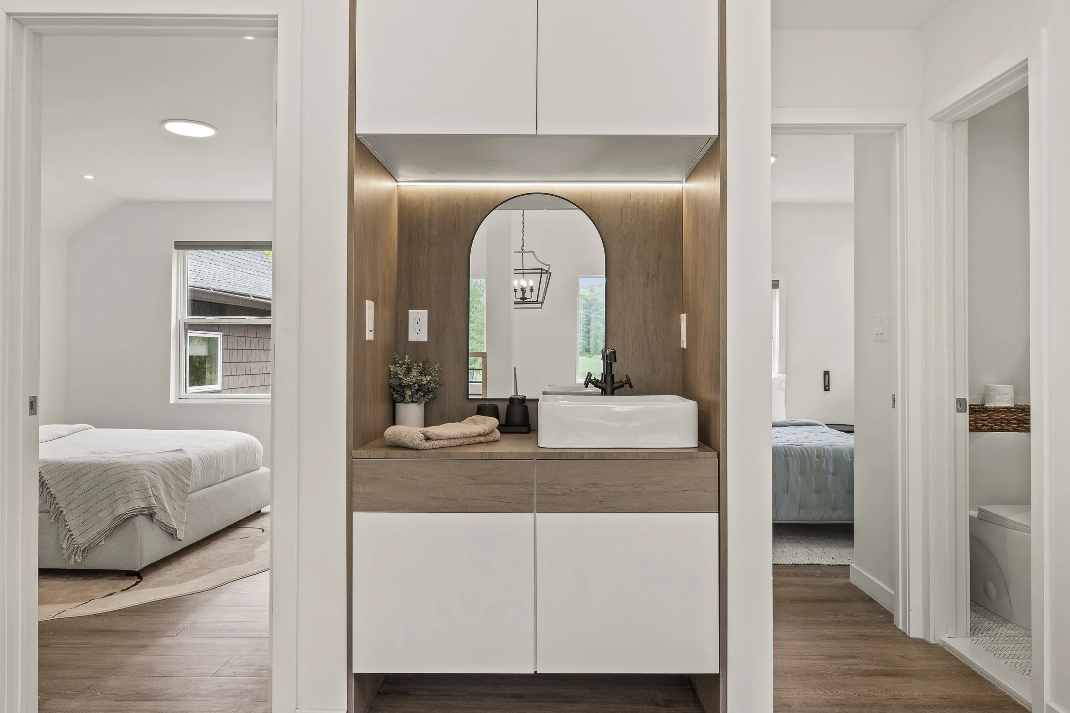 Modern bathroom with a white vessel sink, black faucet, small plant, and beige towels on wooden and white cabinetry, arched mirror, with bedrooms in the background.