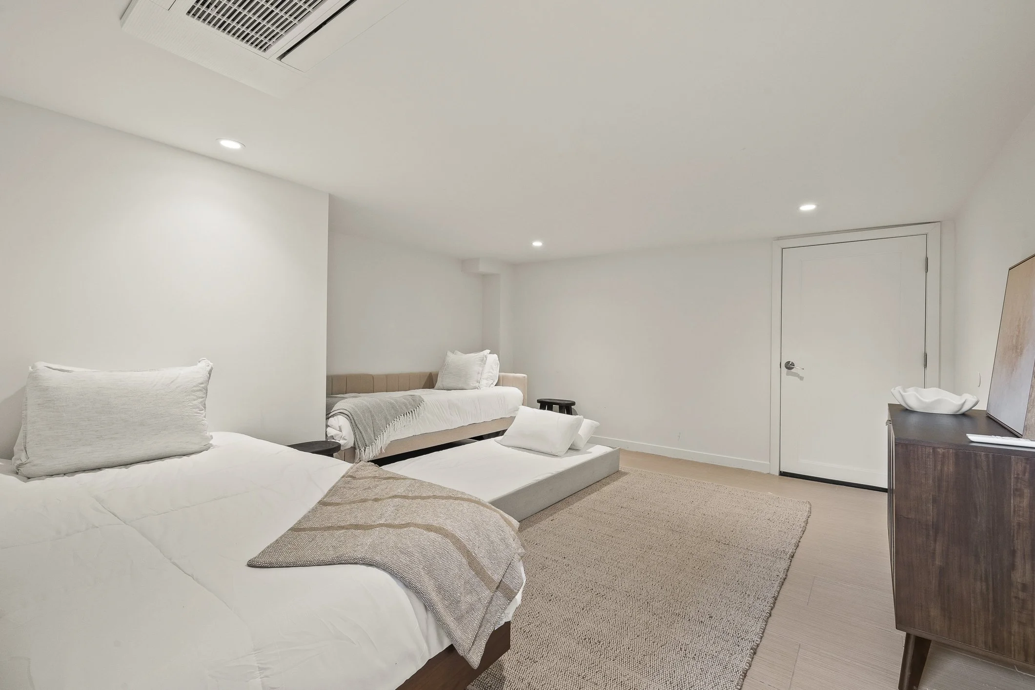 Minimalist bedroom with two beds, a beige bench, a small black side table, a wooden dresser, a beige area rug, and white walls.