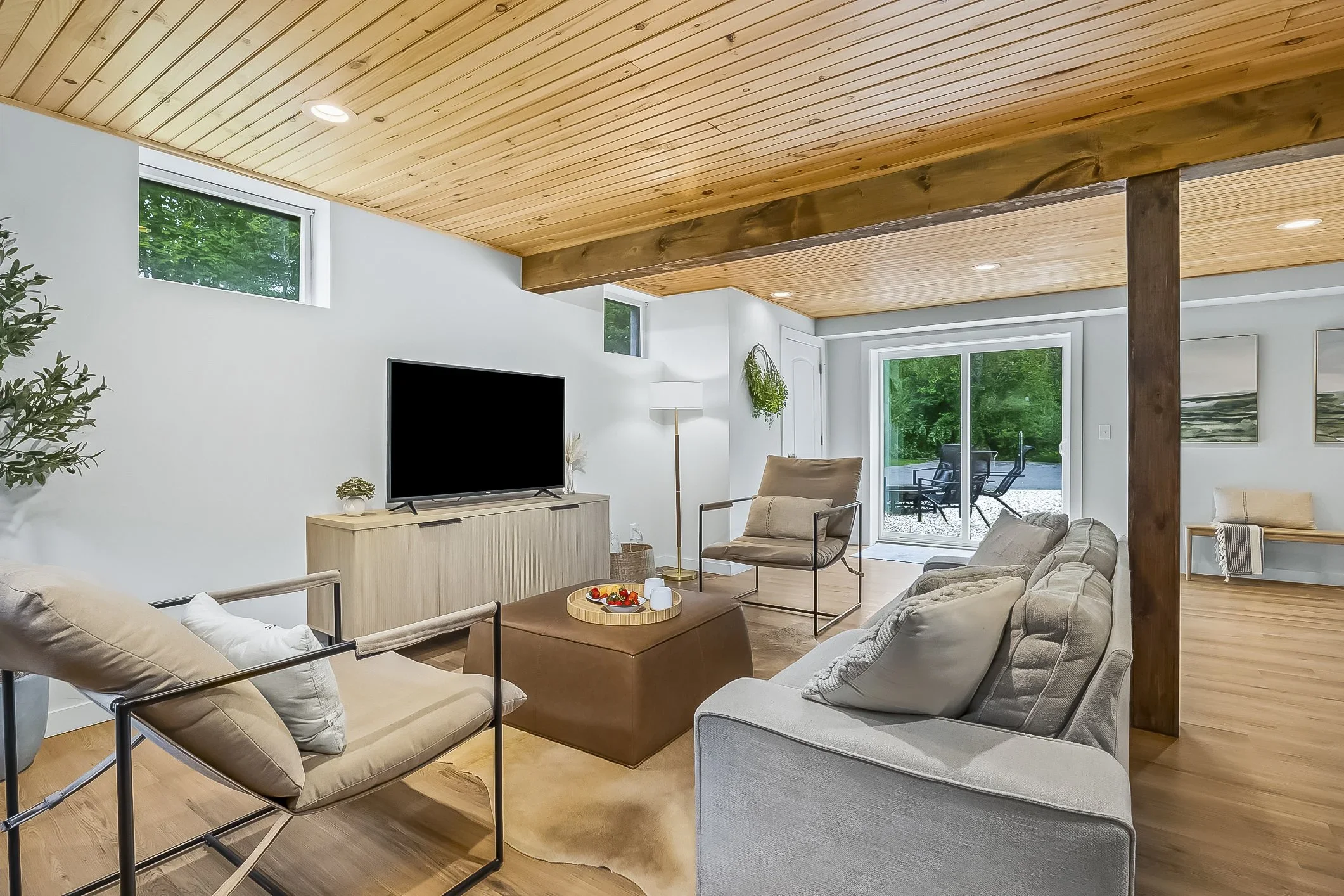 Living room with beige and gray couches, a wooden coffee table with a tray of snacks, a television on a wooden stand, a sliding glass door leading outside, and a wood-paneled ceiling.