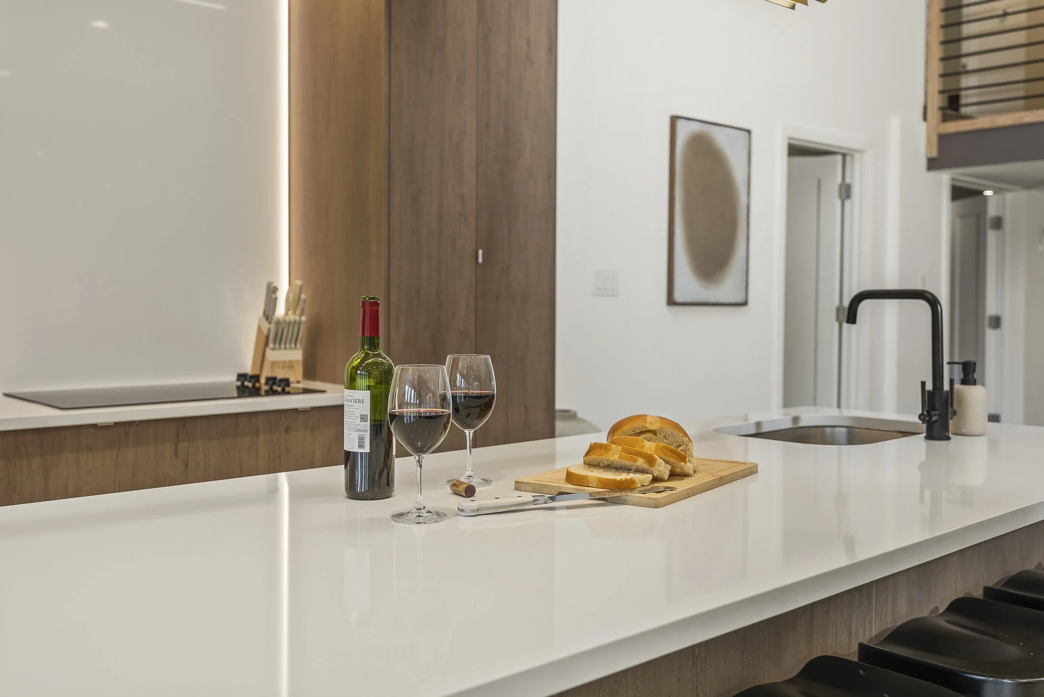 Modern kitchen with white countertop, wine bottle, two glasses of red wine, sliced bread on a wooden cutting board, sink with black faucet, and artwork on the white wall.