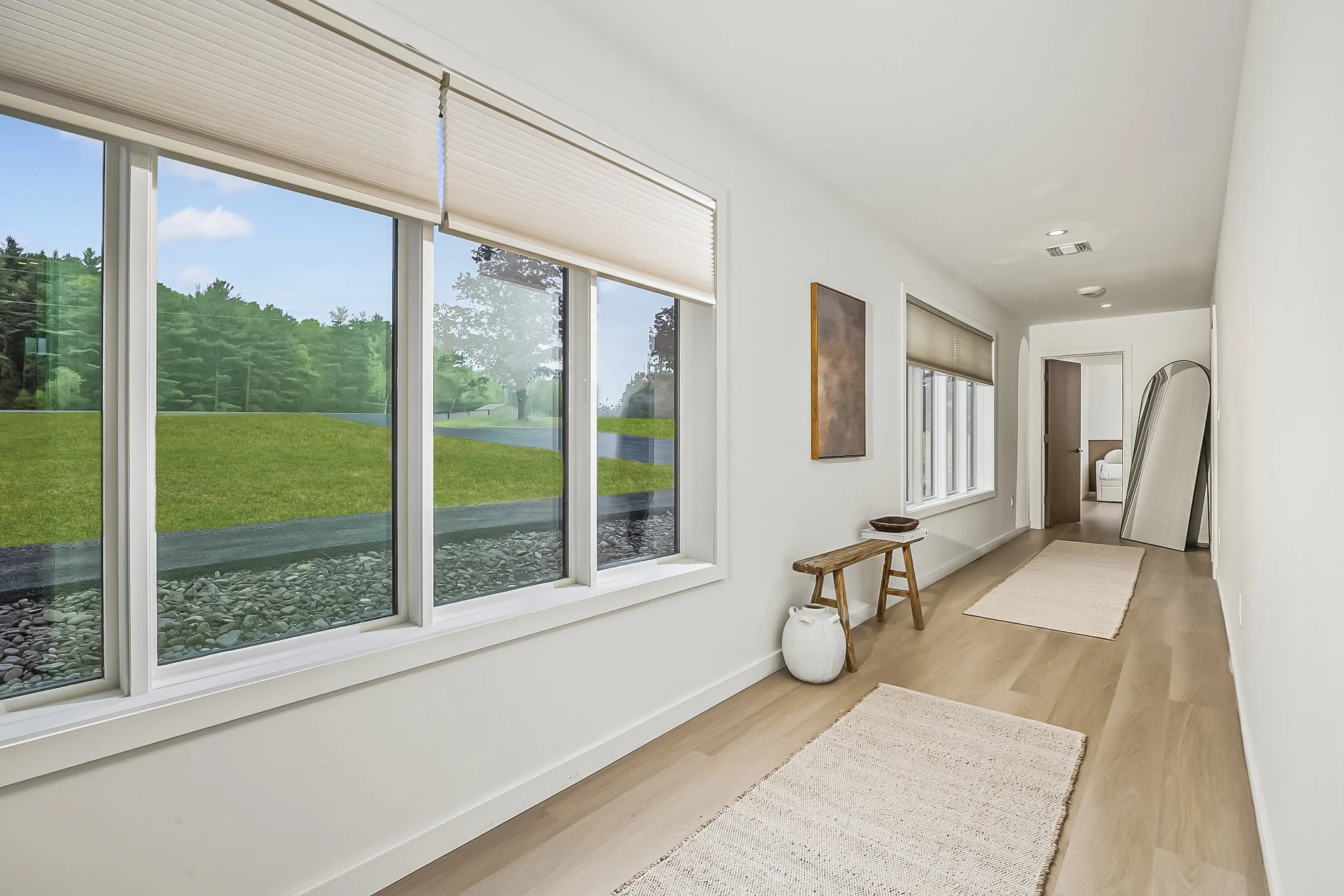 Bright hallway with large windows showing green lawn outside, minimalist decor, wooden bench, artwork, and a mirror leaning against the wall.