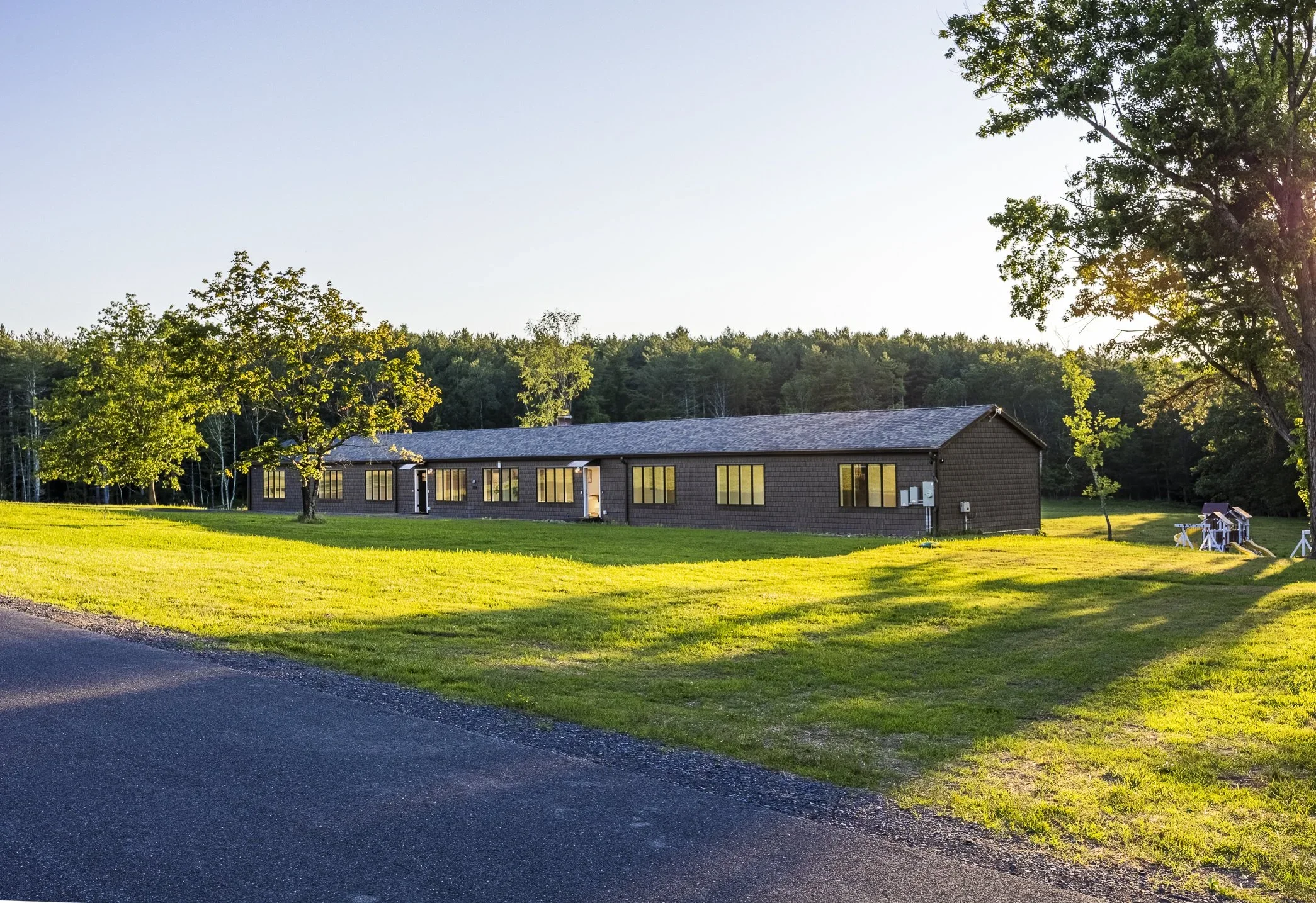 Single-story brown house with multiple windows, surrounded by green grass and trees, with sunlight shining on the scene.