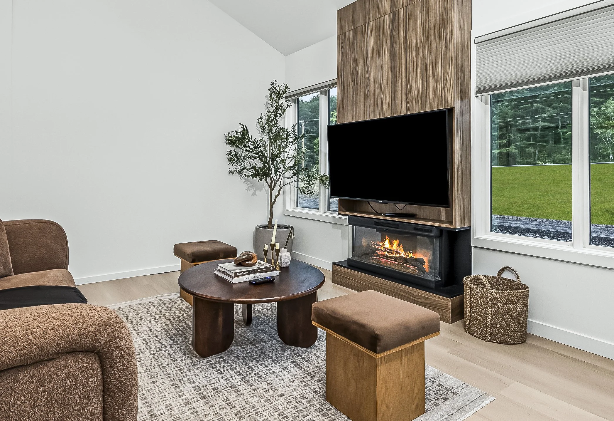 Modern living room with a brown sofa, a wooden coffee table with books and candles, a fireplace, a large flat-screen TV, a potted plant, and large windows revealing a green outdoor landscape.
