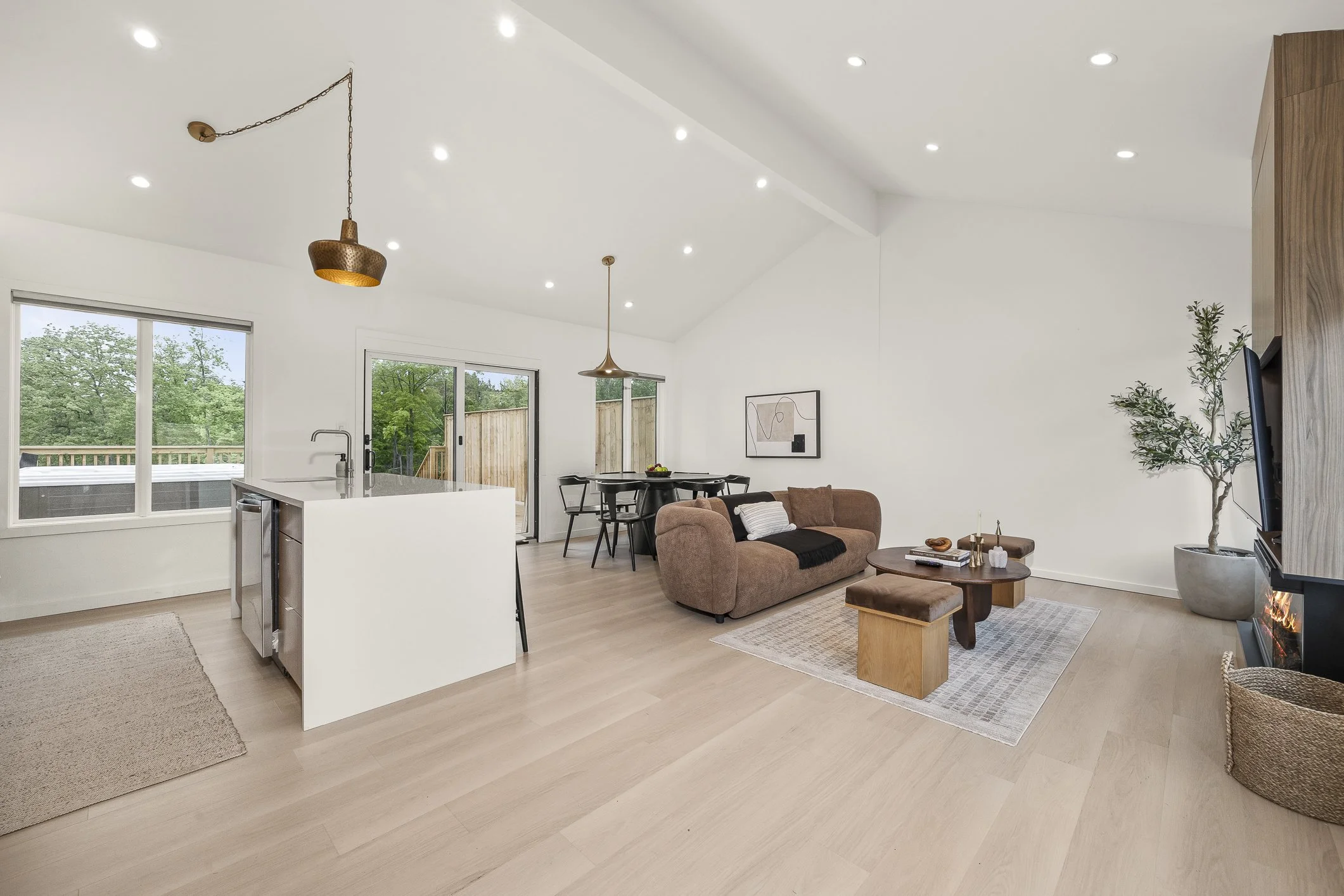 Open-concept living room and kitchen with white walls, light wood floors, brown sofa, coffee table, potted plant, wall art, fireplace, dining area, and large windows showing greenery outside.