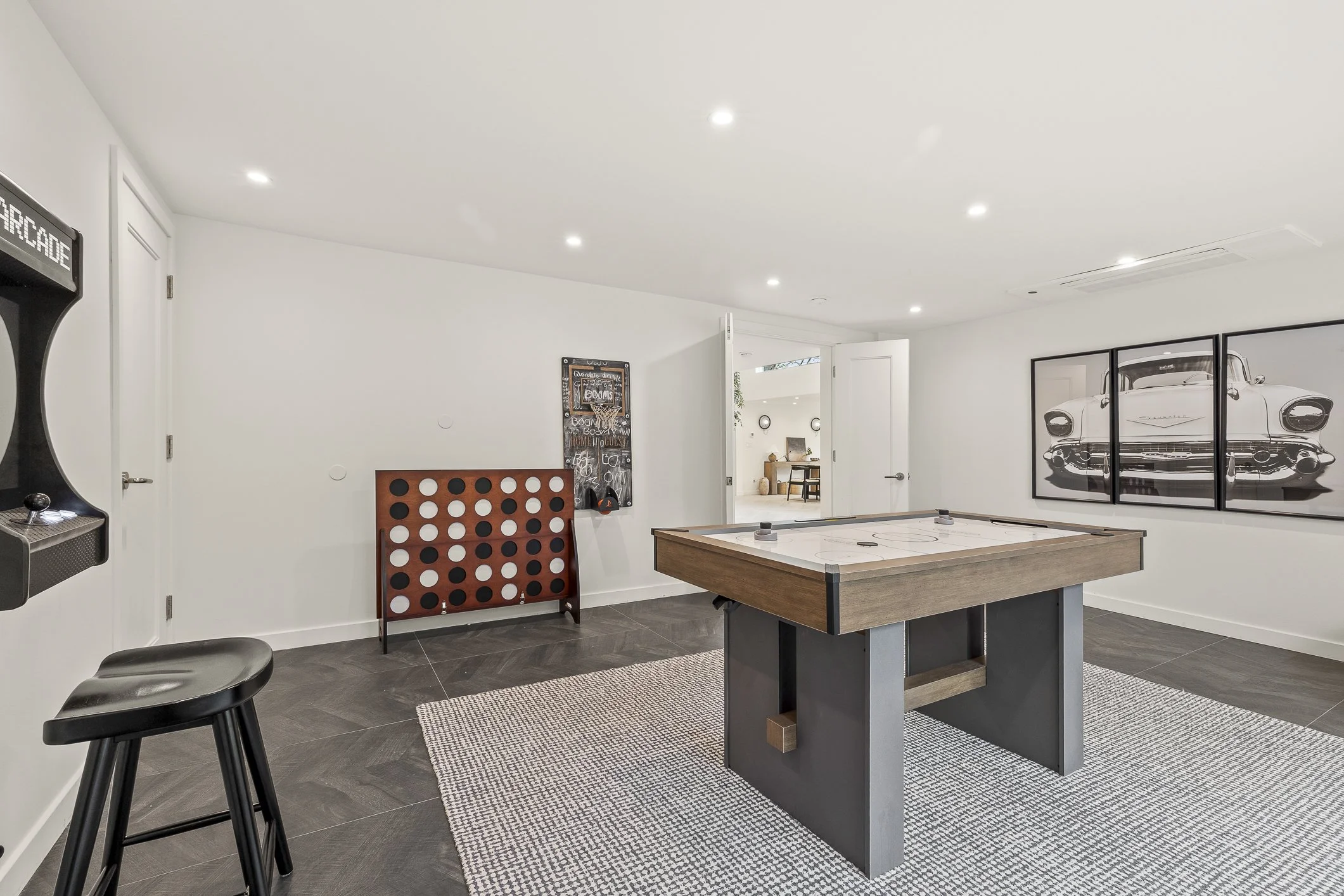 A recreation room featuring an air hockey table, a framed black and white car photo on the wall, a small dartboard, and a vintage arcade game machine, all in a modern home with white walls and dark flooring.