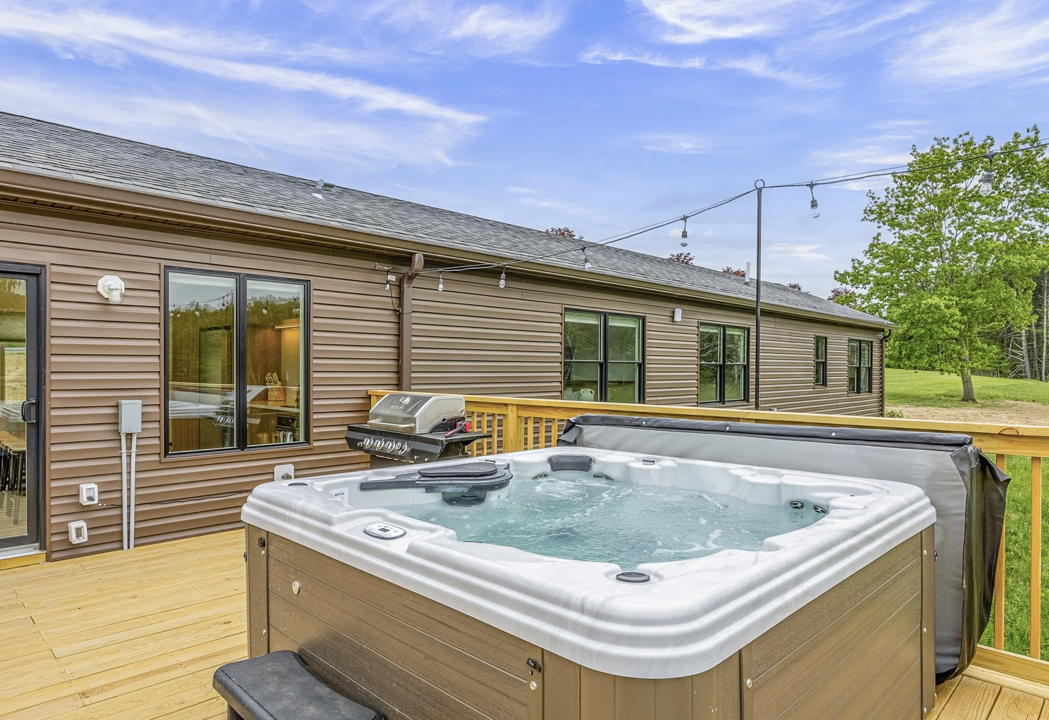Outdoor view of a wooden deck with a hot tub, a grill, and a house with beige siding and multiple windows, under a partly cloudy sky.