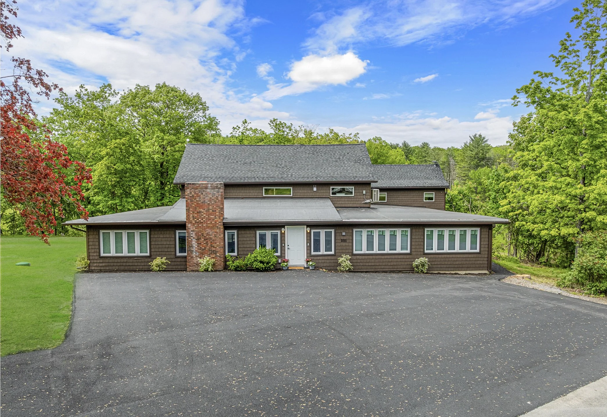 Exterior view of a two-story house with a brick chimney, brown siding, and numerous windows surrounded by green trees under a partly cloudy sky.