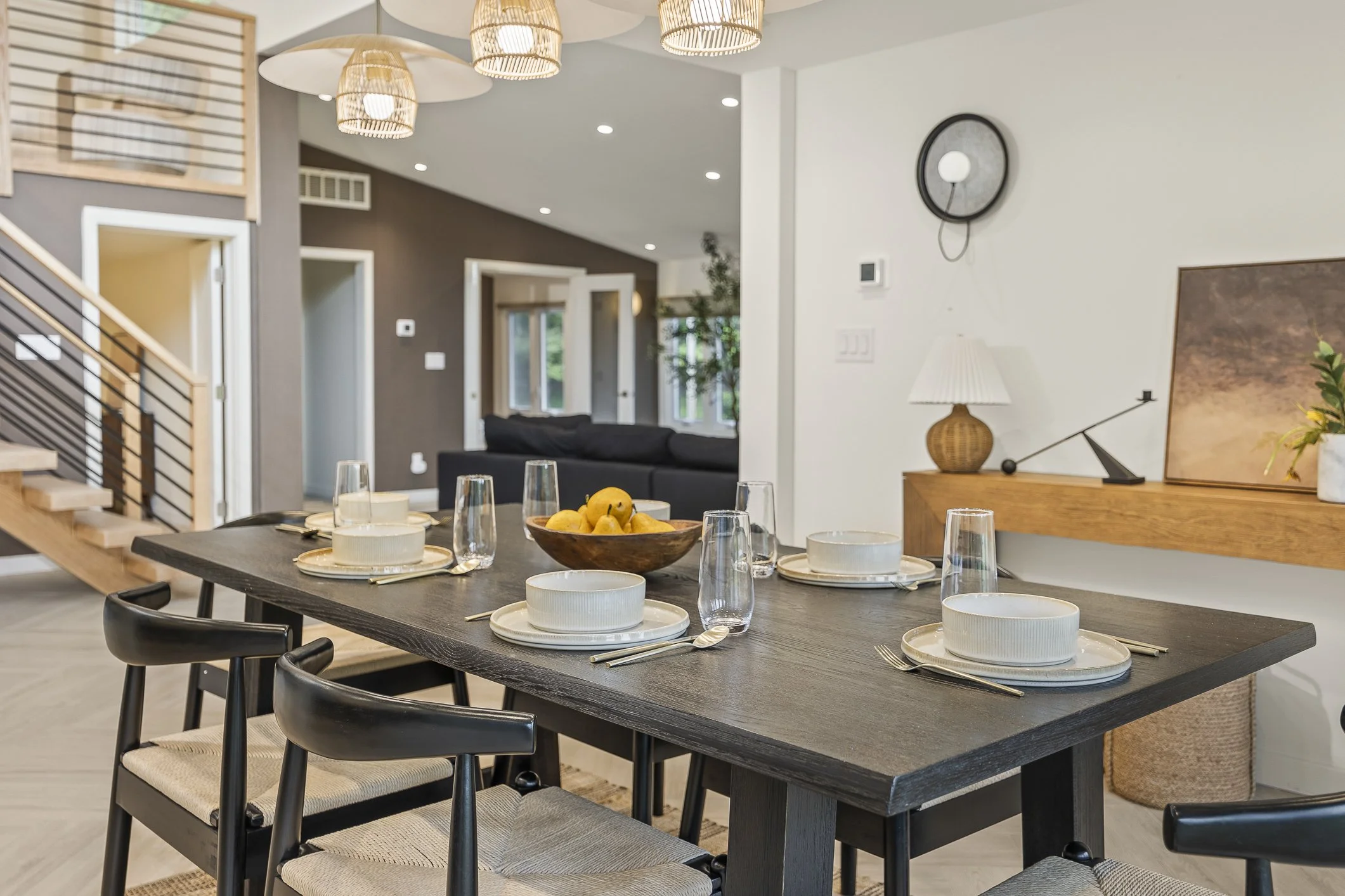Modern dining area with a dark wooden table set for four, featuring white bowls, plates, forks, and glasses, with a bowl of lemons as centerpiece. The background shows a living space with a black sofa, a wall clock, a table lamp, and decorative items