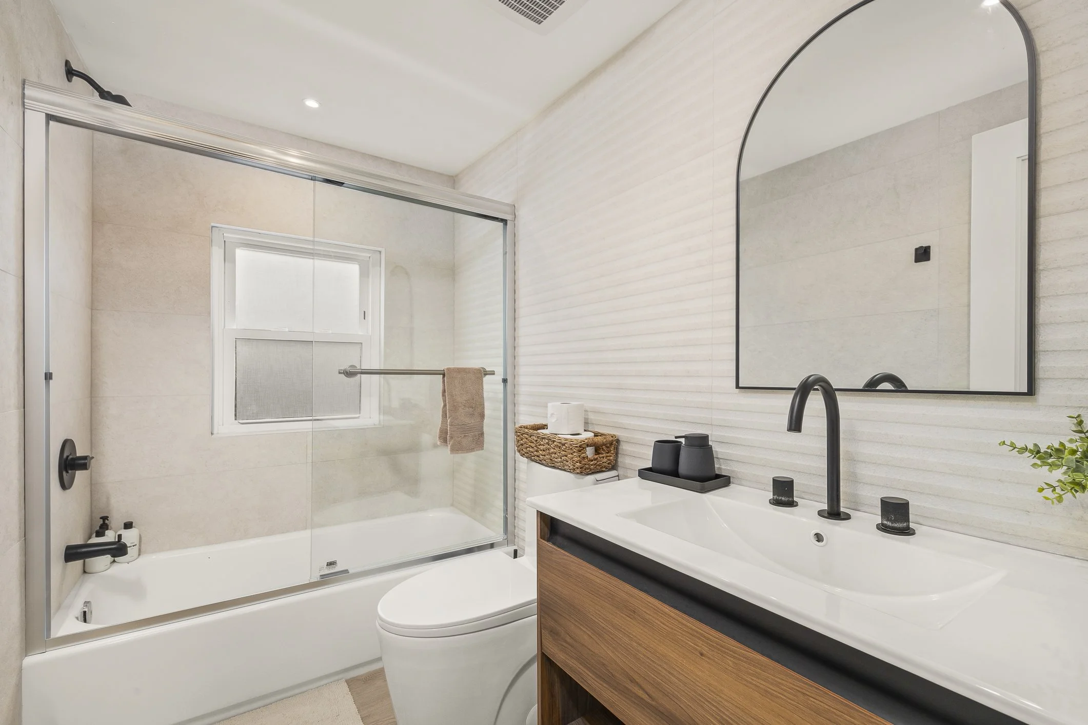 Modern bathroom with a bathtub, a glass shower enclosure, a toilet, and a sink with a large mirror. Neutral tones, black fixtures, and a small window.