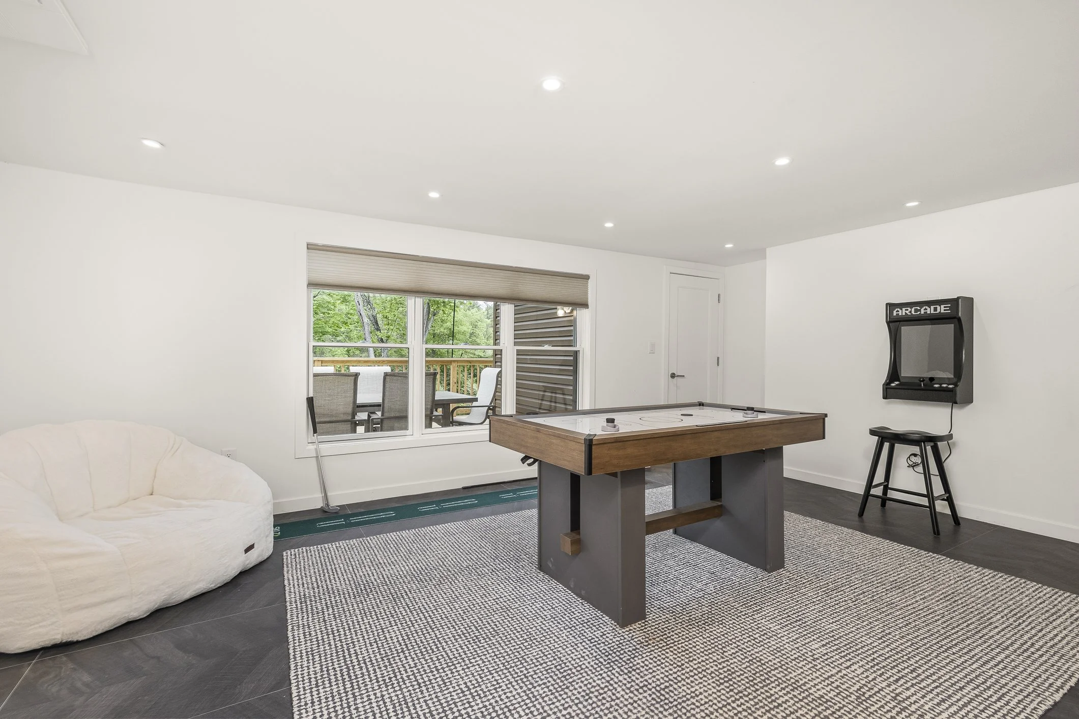 A basement recreation room with white walls and ceiling, dark tile flooring, a white couch, a rug, a dartboard on the wall, and an indoor hockey game table in the center. There is a large window with a view of an outdoor patio with chairs and trees.