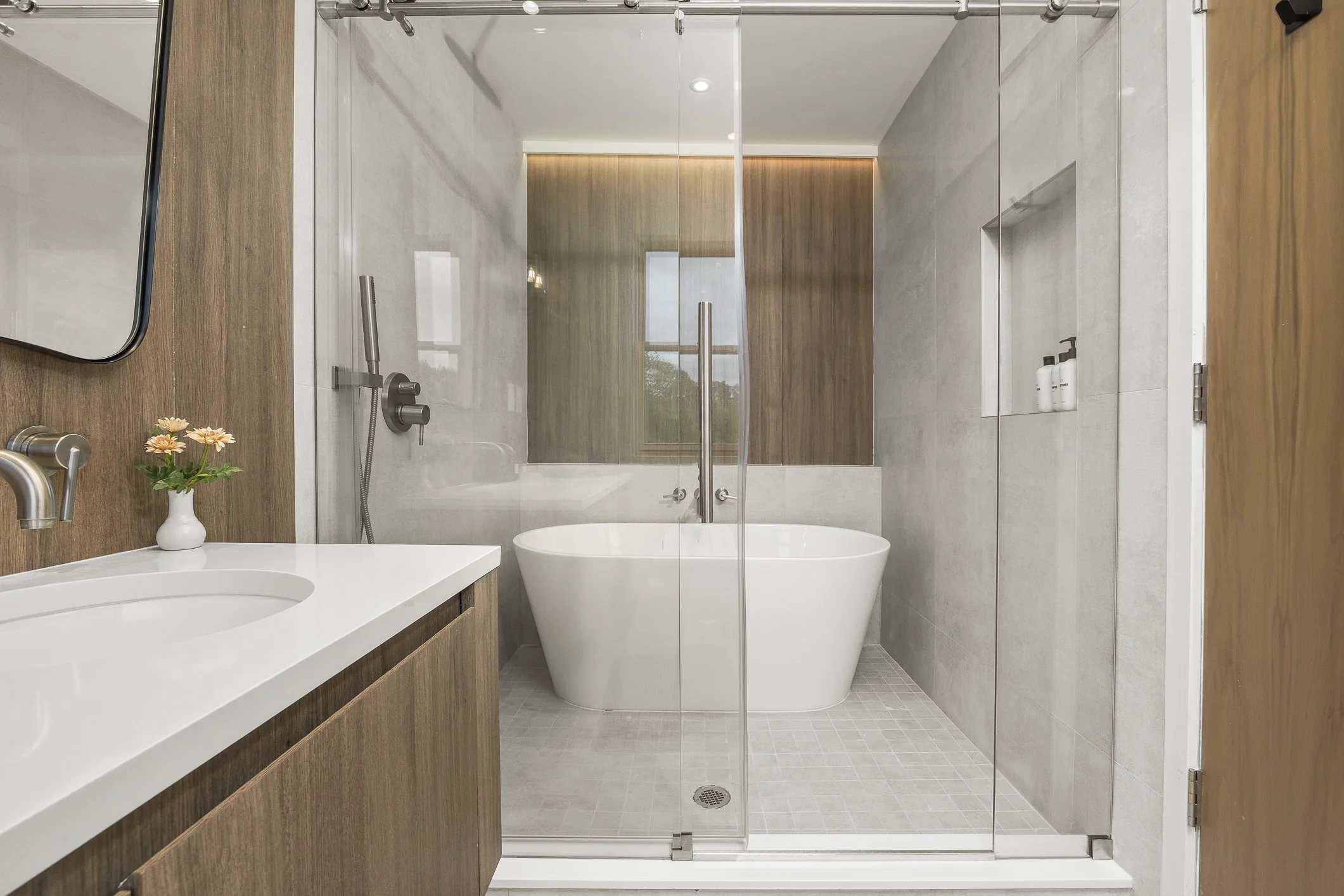 Modern bathroom with a white bathtub, glass shower door, and wood accents, including a vanity with a white sink and a small vase with flowers.
