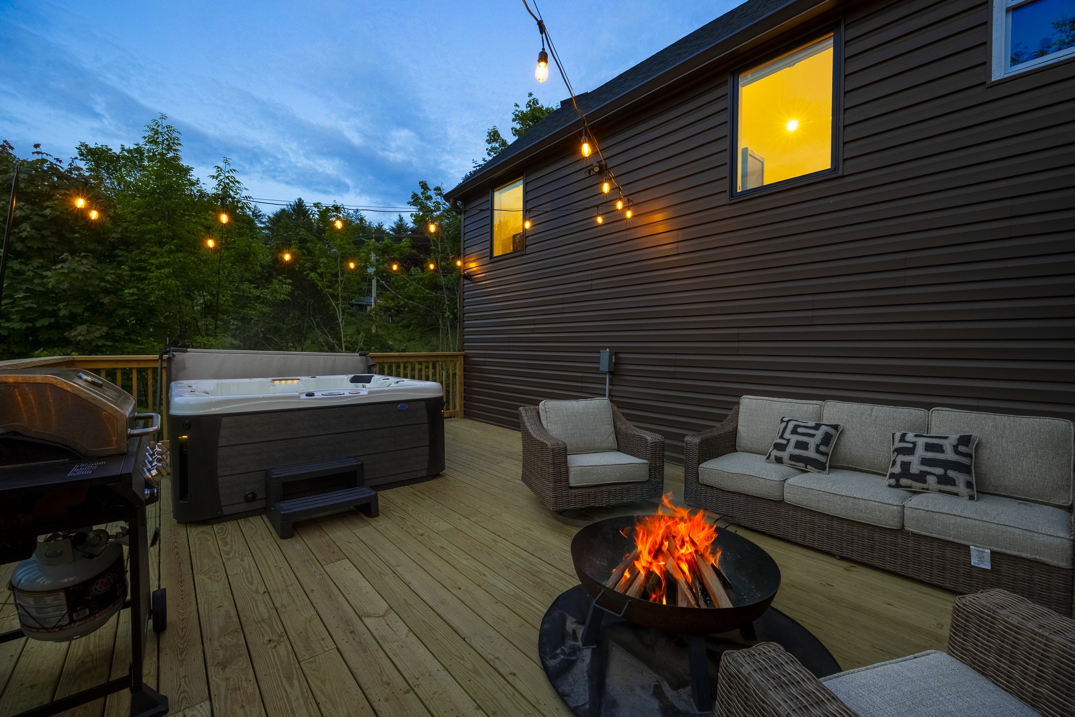A cozy outdoor backyard deck at dusk with string lights, a fire pit, outdoor seating, a hot tub, and a grill, with trees and a house with lit windows in the background.