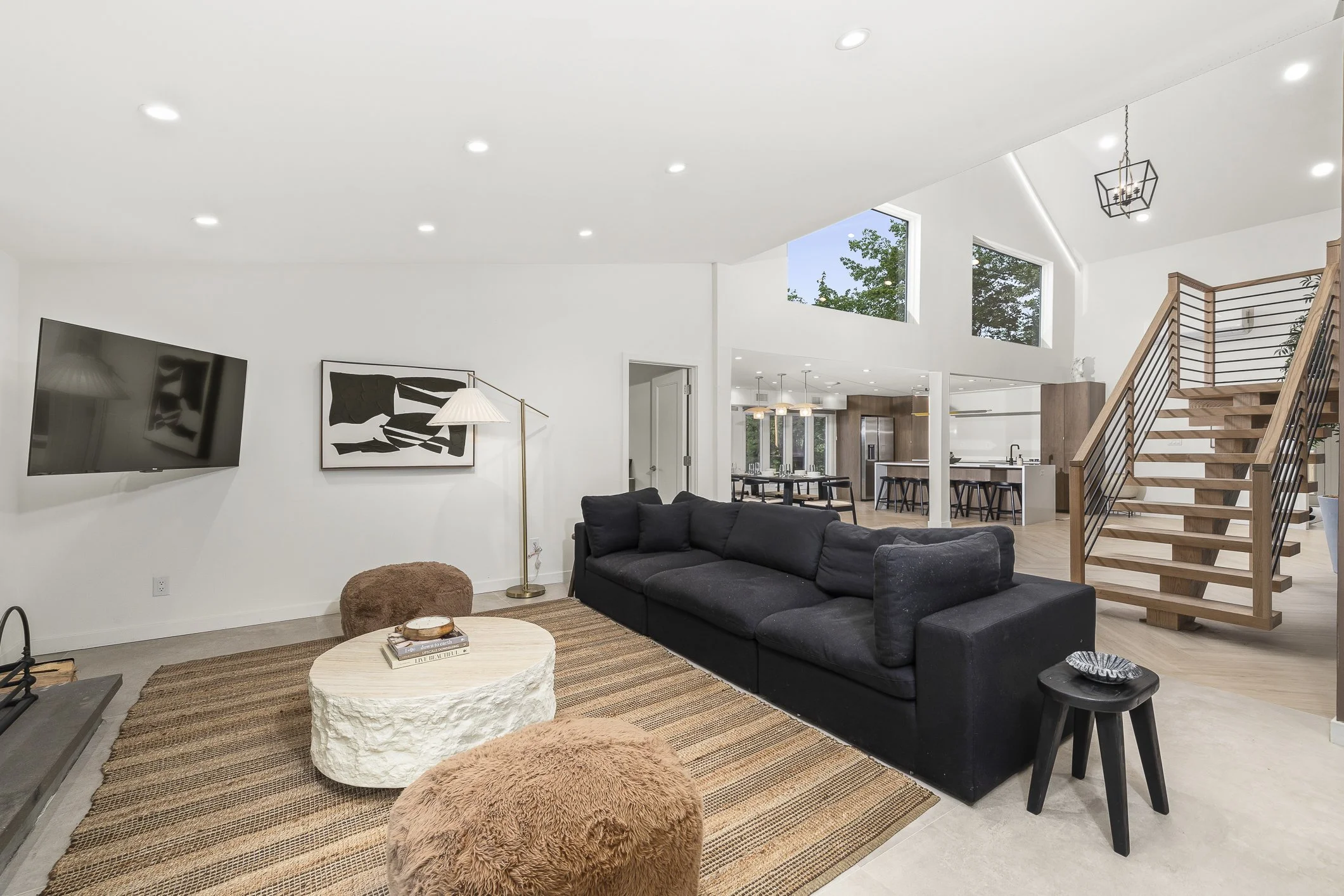 Modern living room with white walls, black sectional sofa, round textured coffee table, brown fuzzy ottomans, flat-screen TV, abstract black-and-white artwork, and a wooden staircase with metal railing leading upstairs. Open-concept area with kitchen