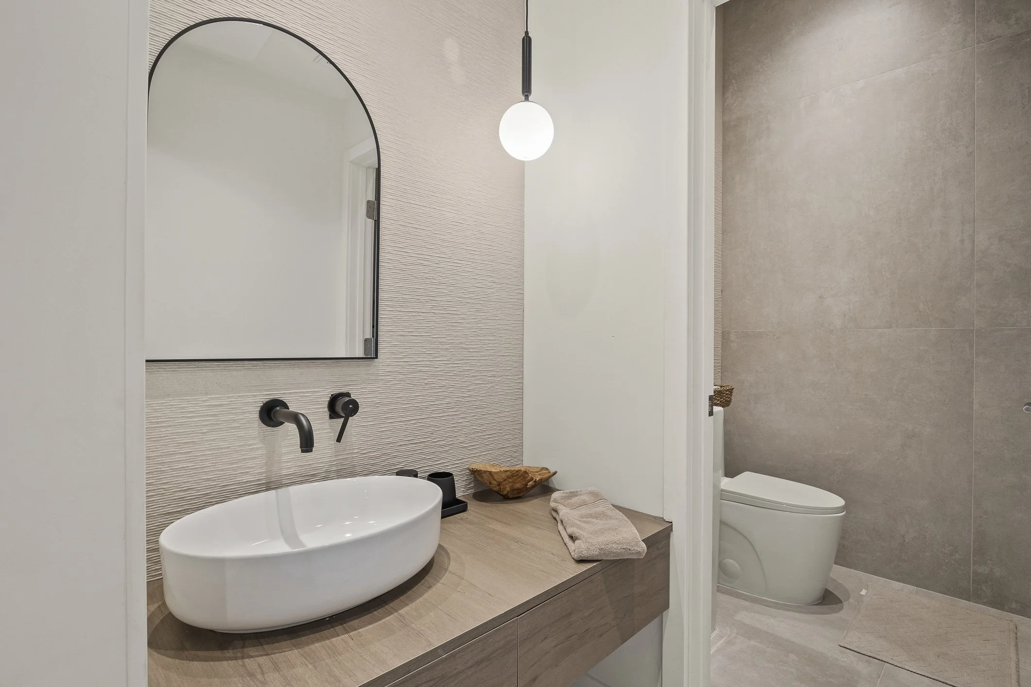 Modern bathroom with a floating wooden vanity, oval sink, black wall-mounted faucet, a mirror, a hanging light, a beige towel on the vanity, and a separate toilet area with beige tiled walls and a small beige rug.