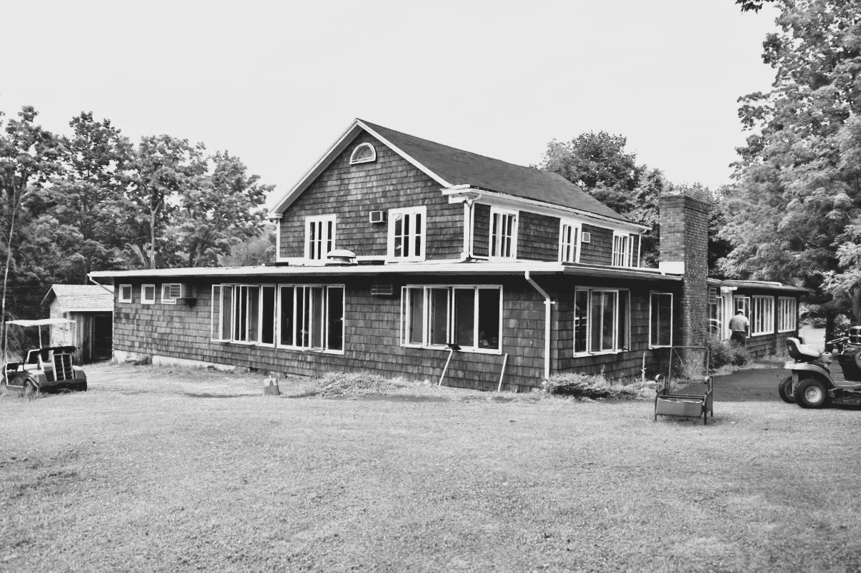 A two-story house with shingle siding, multiple windows, a brick chimney, and outdoor equipment in the yard, surrounded by trees.