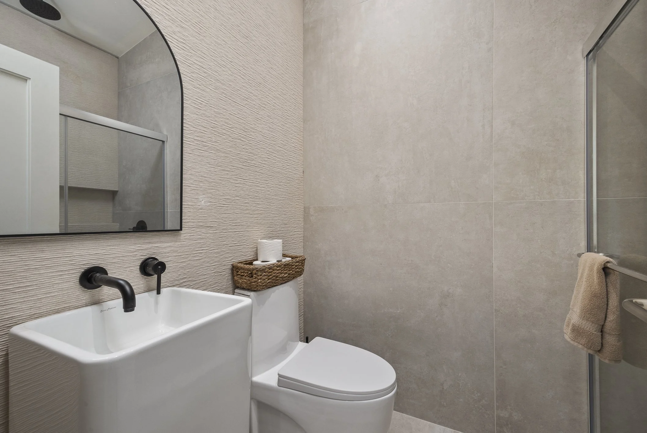 Modern bathroom with beige tile walls, a white rectangular sink with black fixtures, a mirror, a toilet with a wicker basket holding toilet paper on top, and a glass-enclosed shower with a beige towel hanging on the door.