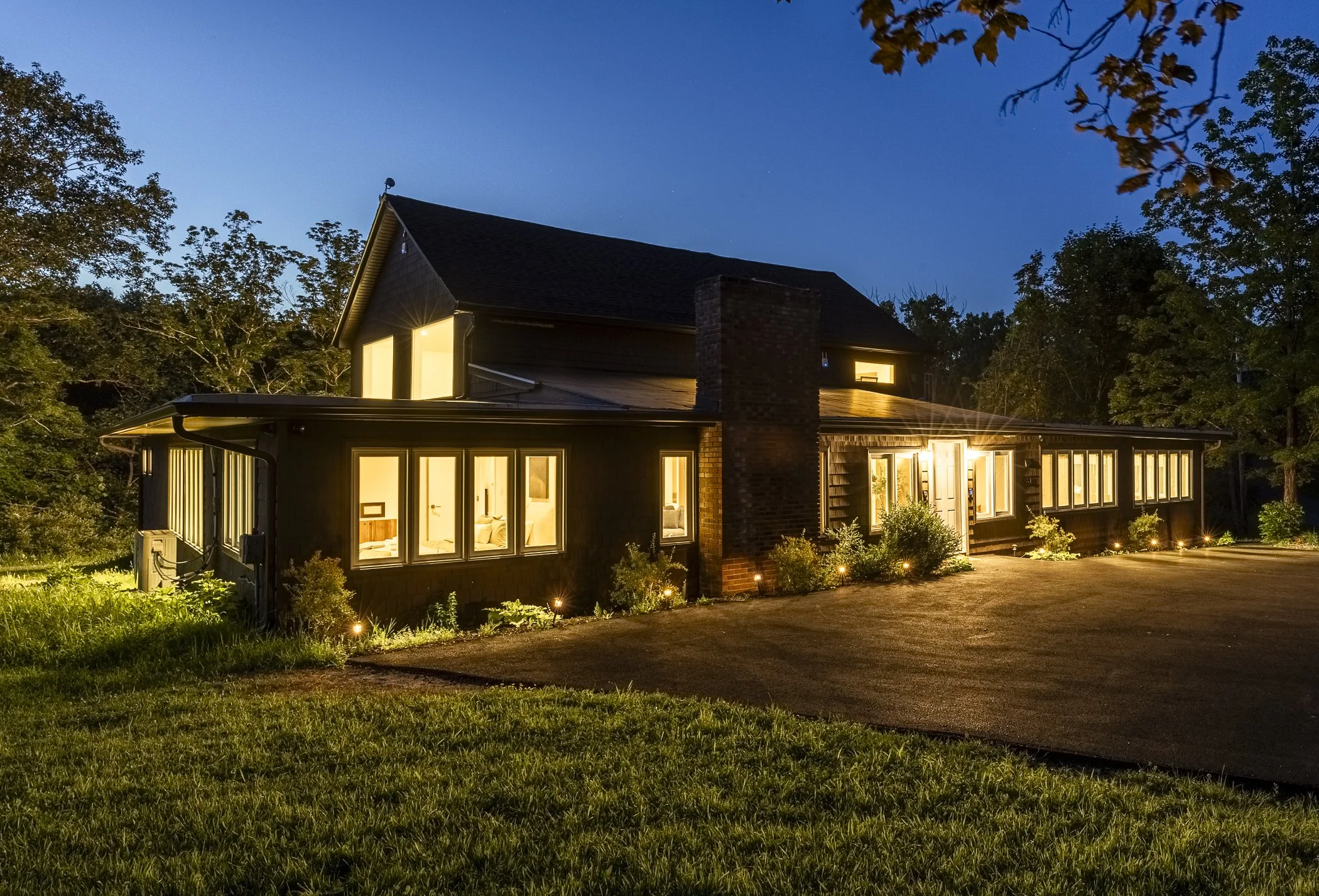 A two-story house at dusk with illuminated windows, brick chimney, surrounded by trees and a grassy lawn, with outdoor lighting along the walkway.