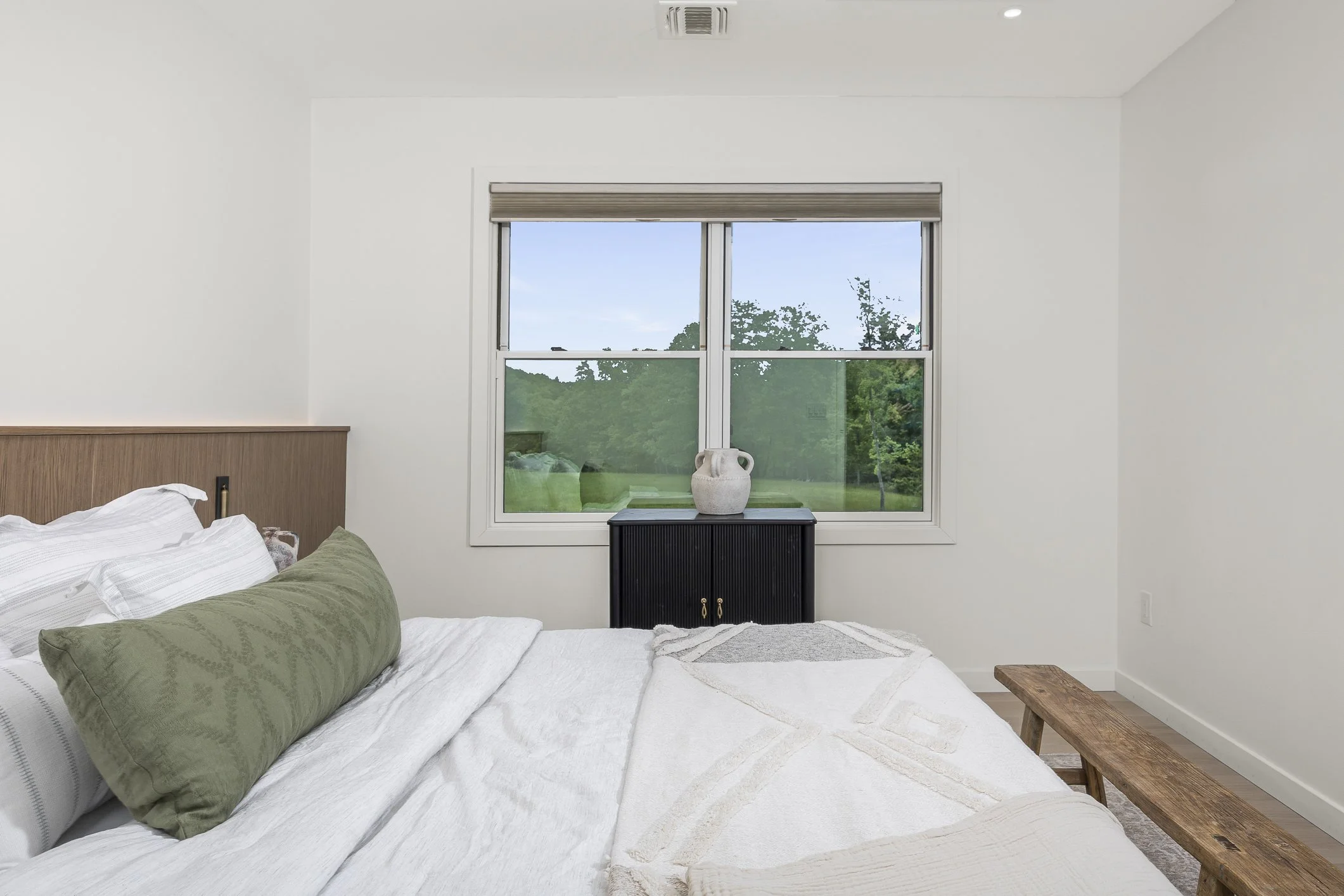 Bedroom with white walls and bed, green pillow, black cabinet, white pitcher, and window showing a green tree and grass outside.
