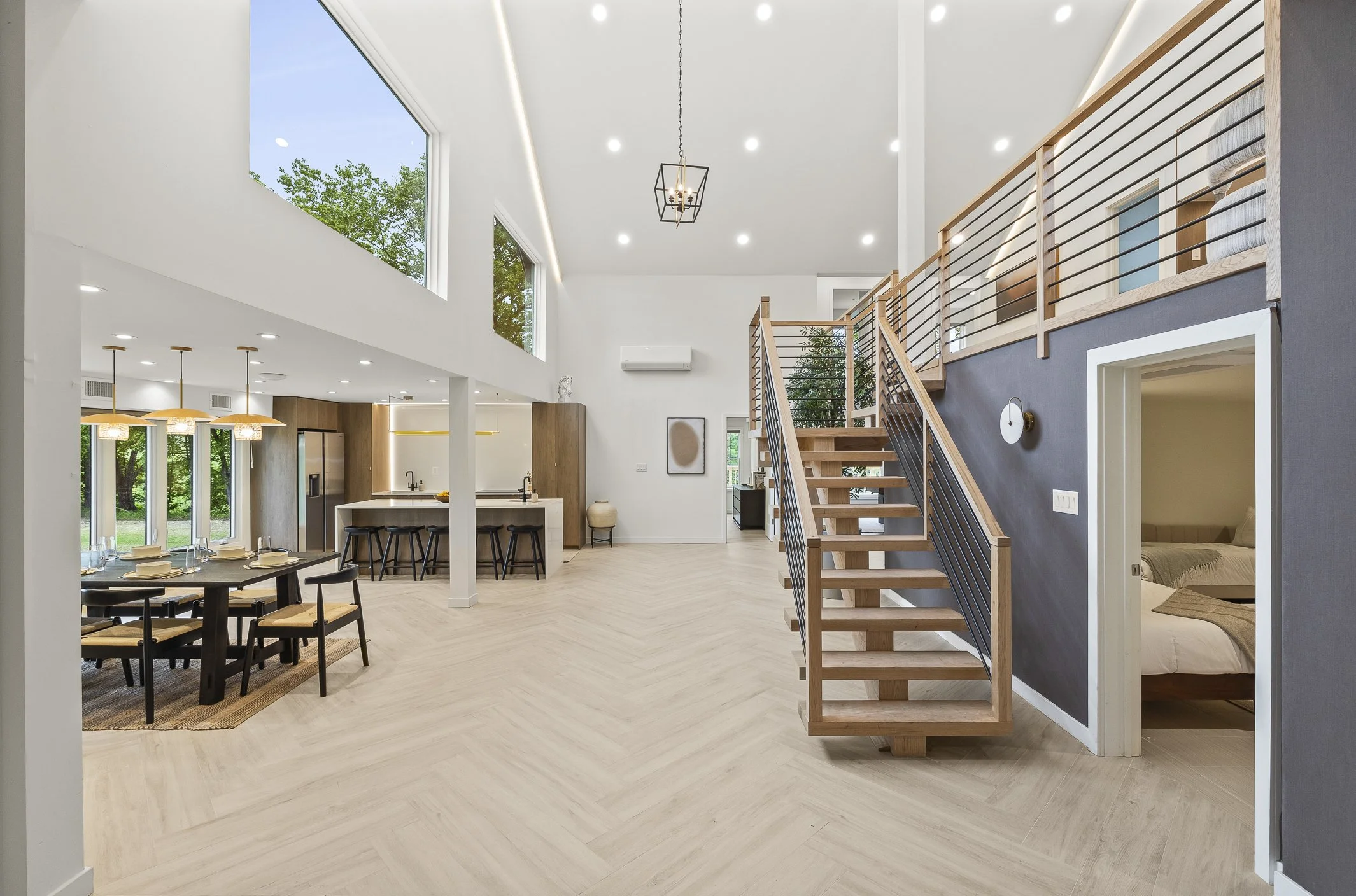 Interior of a modern, spacious open-concept living space with high ceilings, large windows, a kitchen with a breakfast bar, a dining area with a table and chairs, a staircase with wooden steps and black railing, and a visible bedroom through an open 