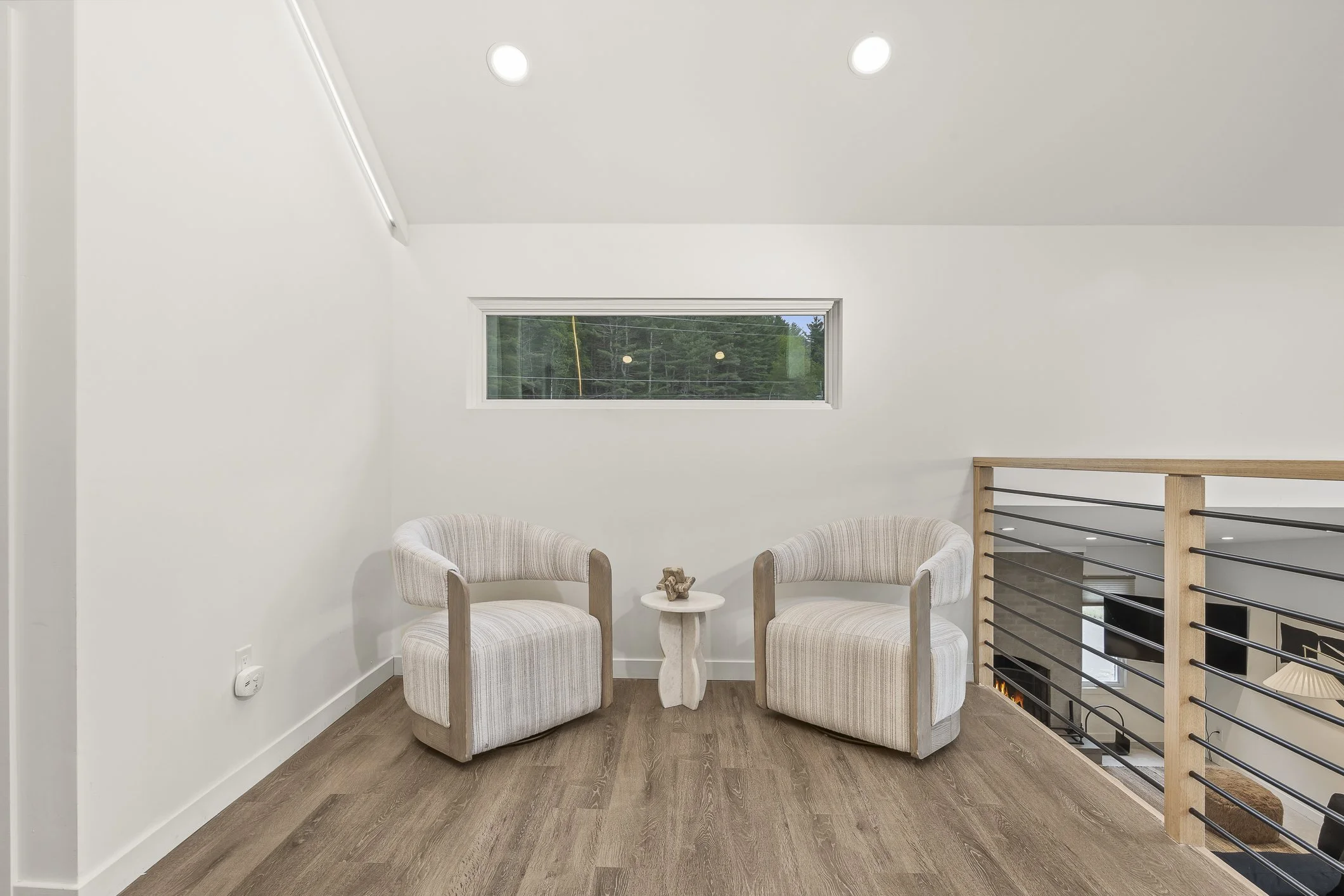 A small seating area with two cream-colored upholstered armchairs, a small white side table, a narrow horizontal window on a white wall, and a wooden railing overlooking a lower level.