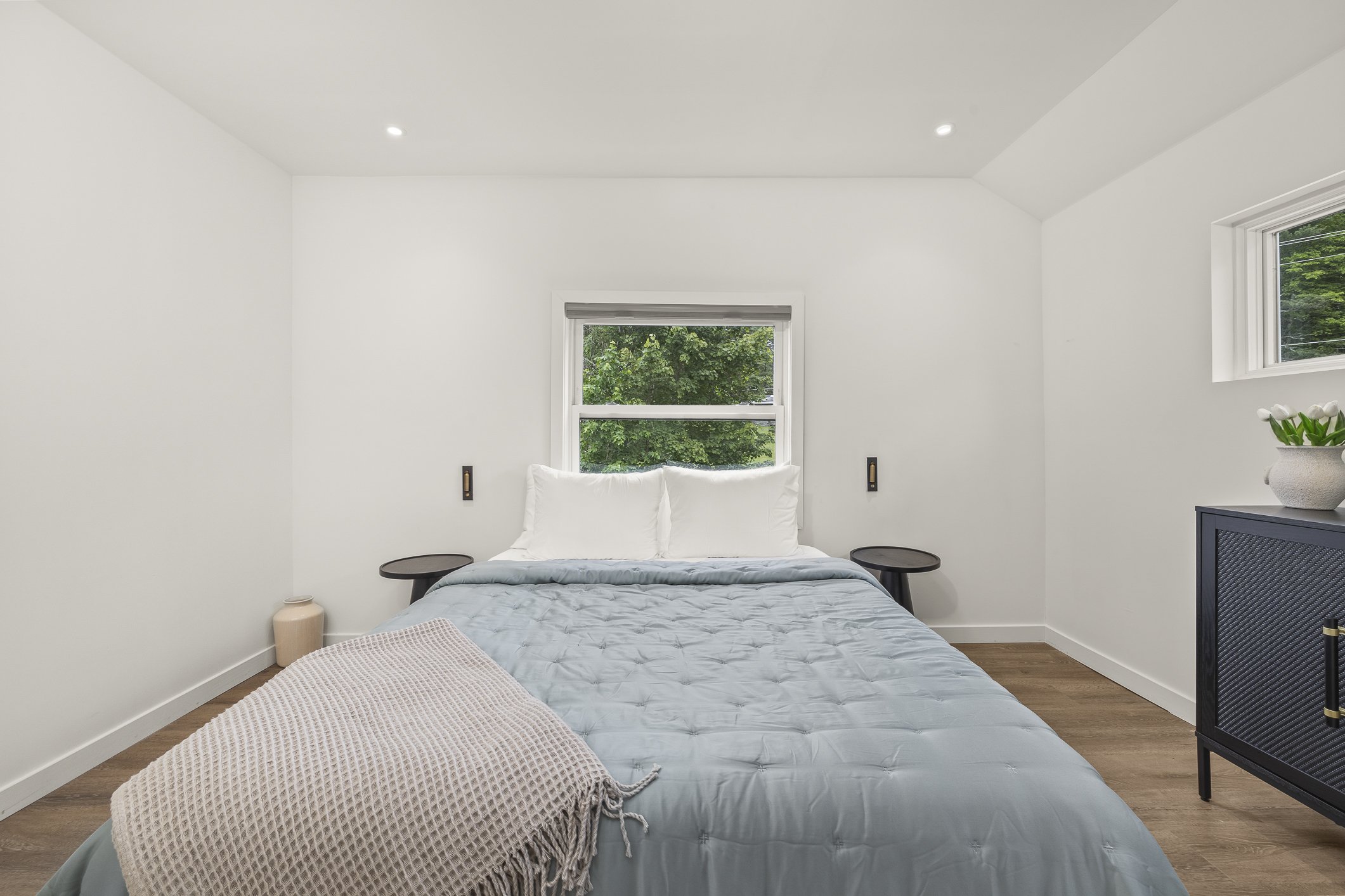 A minimalist bedroom with white walls, a bed with white pillows and light blue bedding, a window with a view of green trees, two black nightstands, a blue cabinet with a white vase of flowers, and a beige throw blanket on the bed.