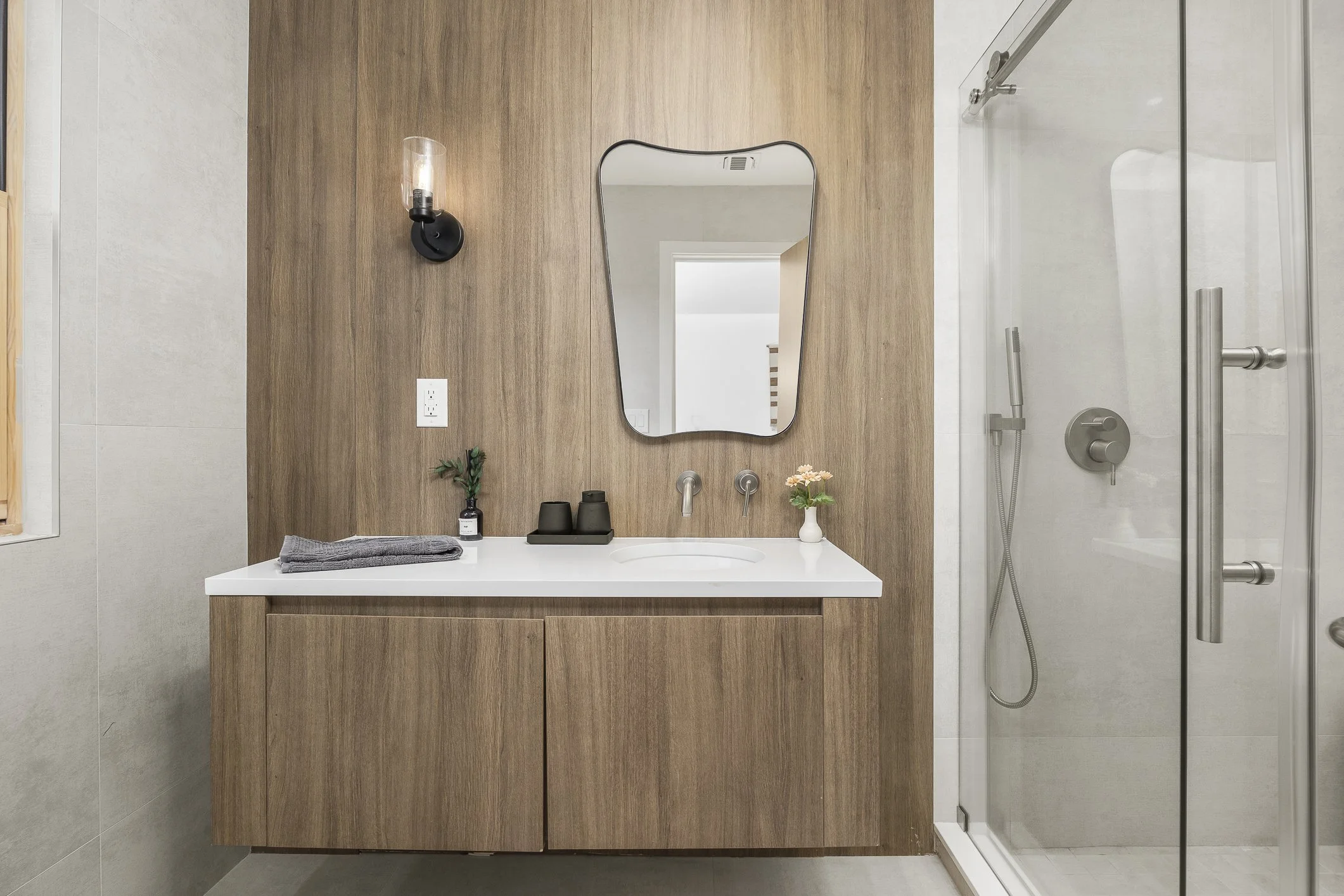 Modern bathroom with a wooden vanity, white countertop, round sink, mirror, black wall sconce, small plant, towel, and a shower with glass door and silver fixtures.