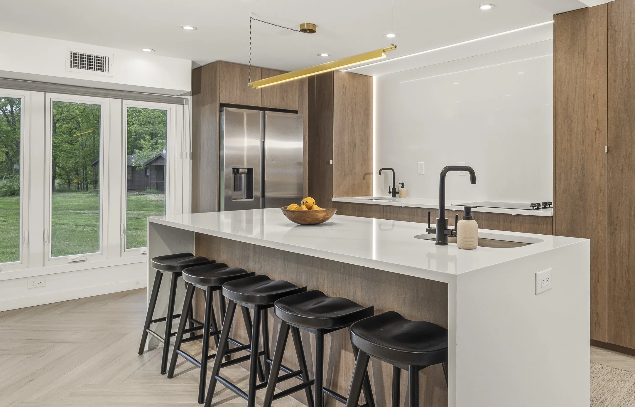 Modern kitchen with white island, black stools, wooden cabinetry, stainless steel refrigerator, and large windows overlooking a green yard.