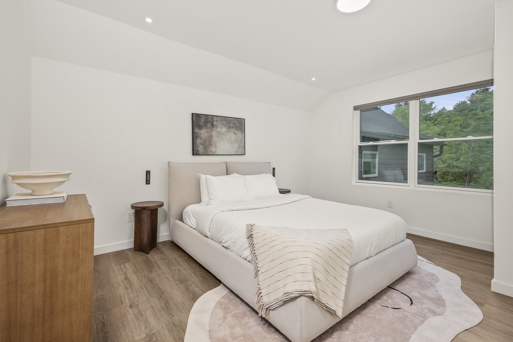 A modern bedroom with a white bed, beige headboard, and white bedding. A tan blanket is draped over the end of the bed. There is a wooden nightstand on each side of the bed, with a dark block-shaped lamp on each. A large window with a view of trees a