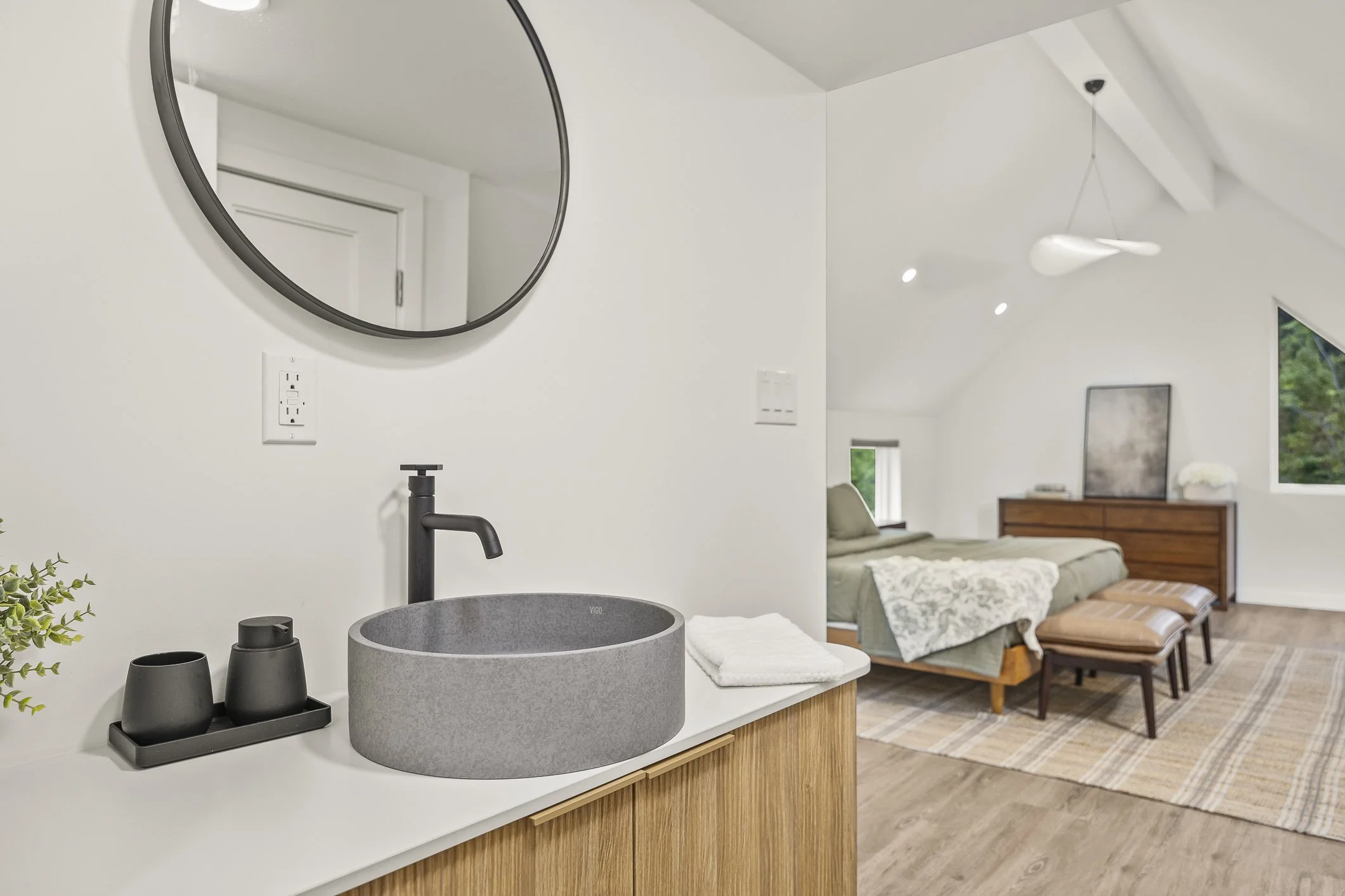 Modern bedroom with a bed, wooden dresser, windows, and a sloped ceiling, seen from a corner with a modern sink and mirror in the foreground.