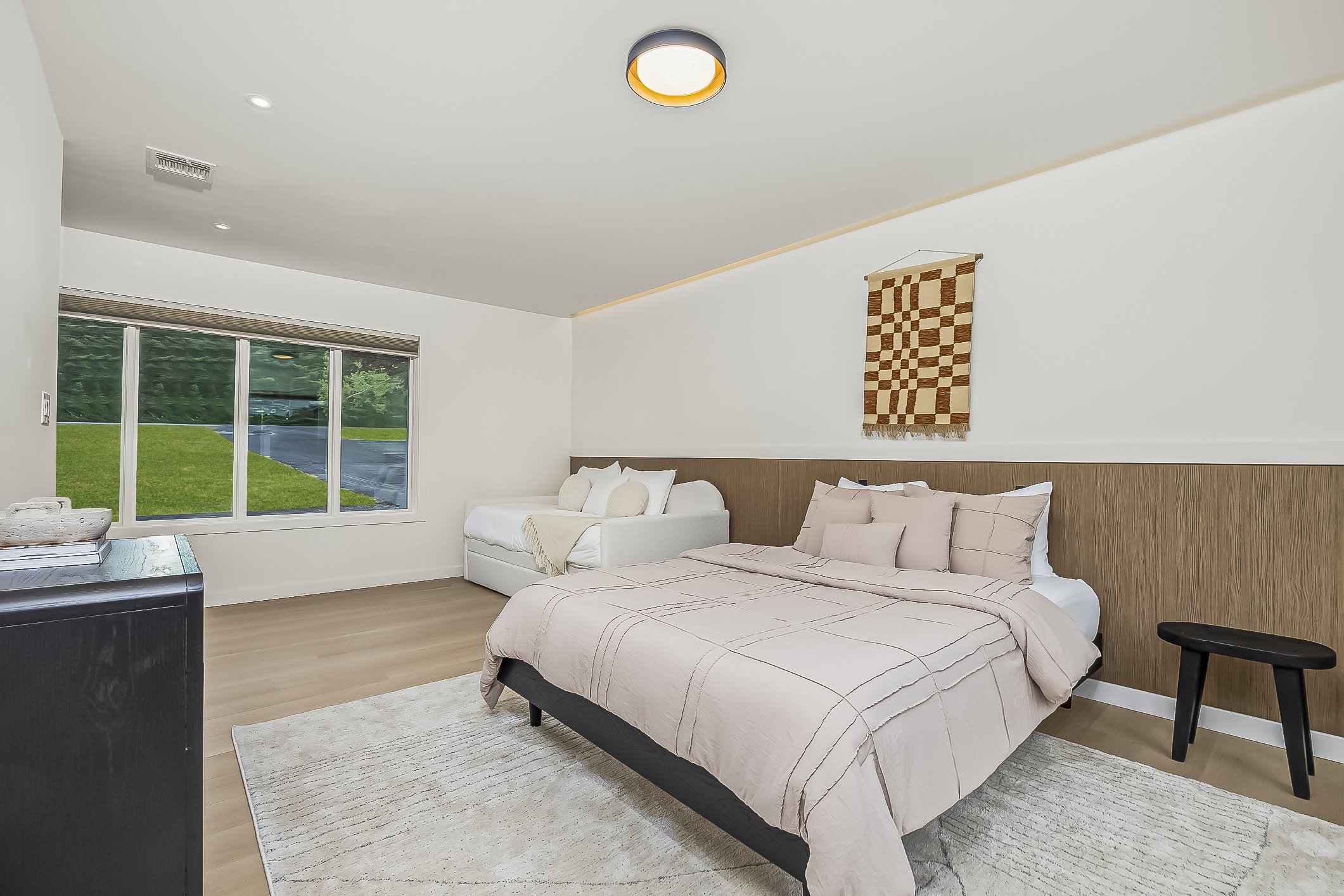 A bedroom with a large bed, a smaller white sofa, a window showing a green outdoor area, a black bedside table, and a geometric wall hanging.