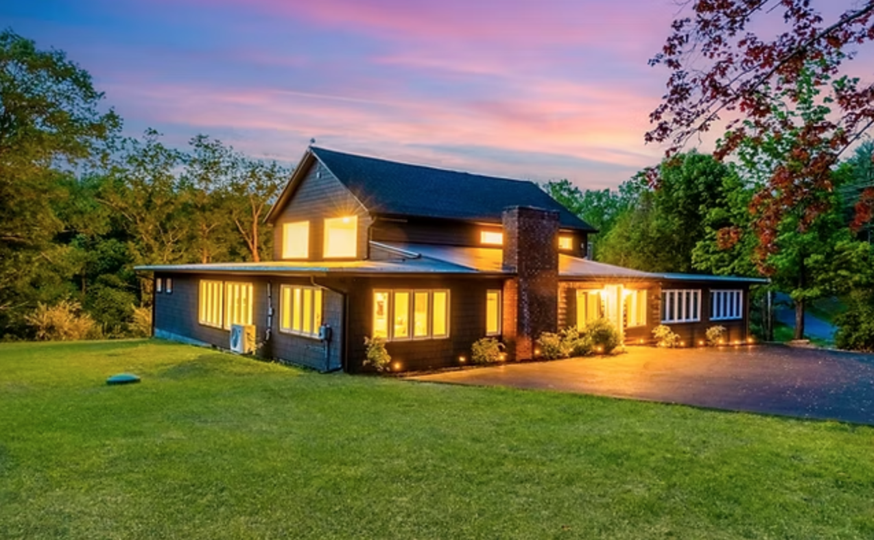 A two-story house with black exterior and yellow-lit windows at dusk surrounded by greenery and a well-maintained lawn.