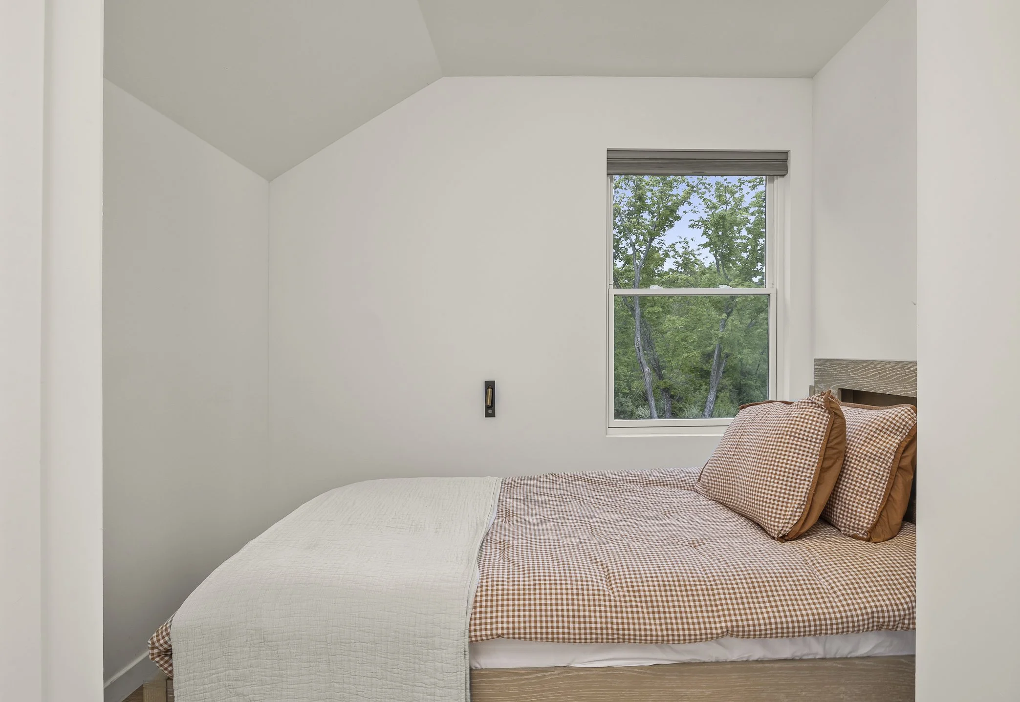 A bedroom with a bed made with orange and white checkered bedding and matching pillows, a white quilt at the foot of the bed, a window showing green trees outside, and a wall outlet on the wall behind the bed.
