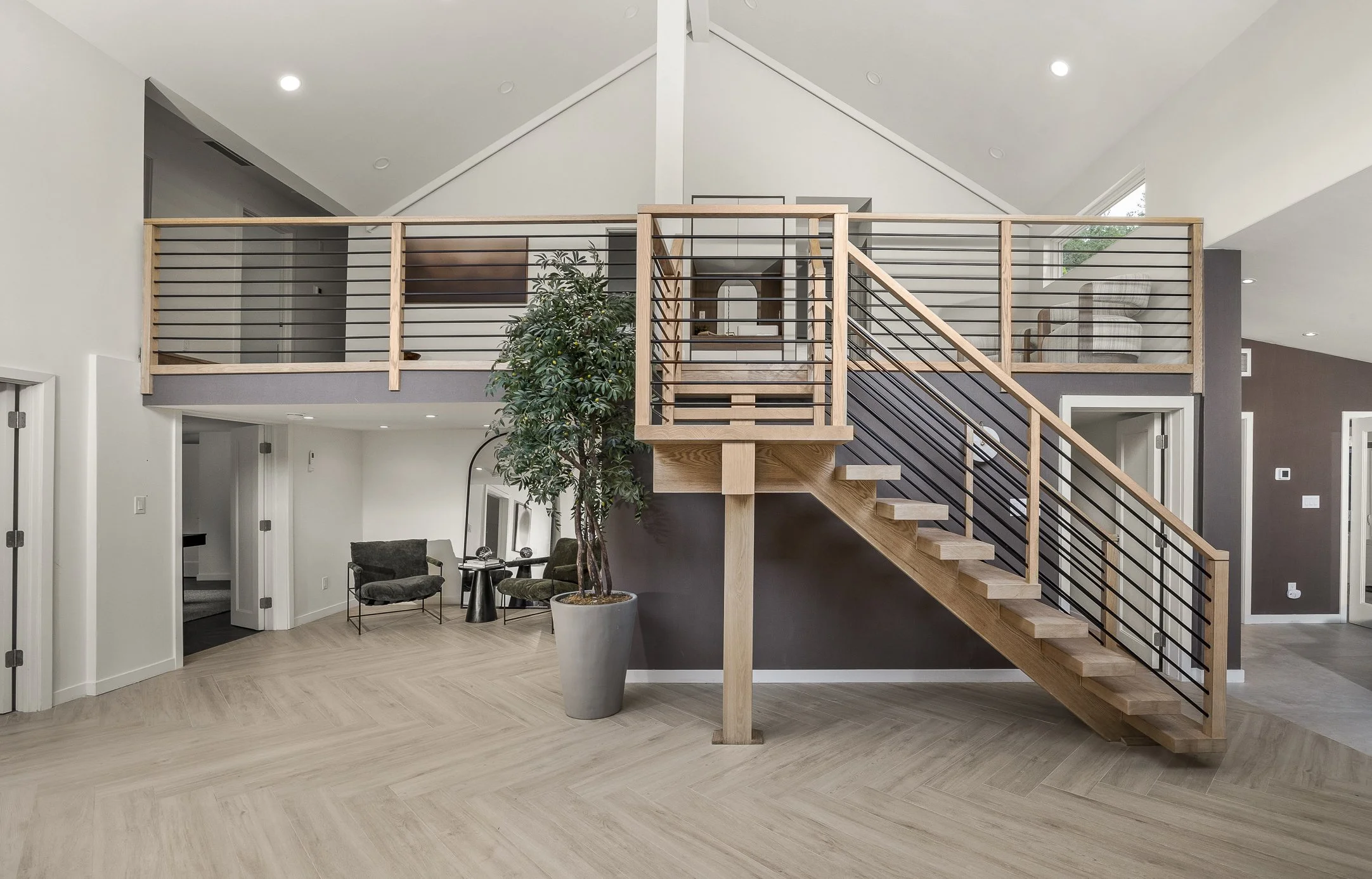 Living room with wooden staircase leading to second-floor balcony, potted plant, seating area, and modern decor.