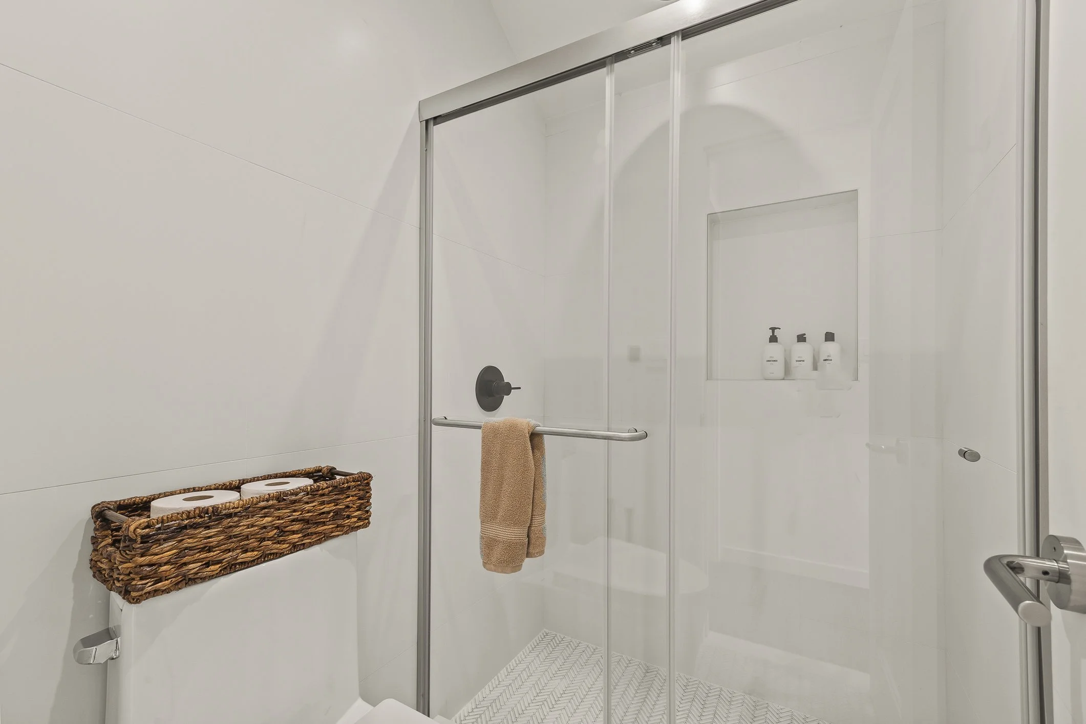 Modern walk-in shower with glass doors, built-in wall niche with toiletries, and a toilet with a woven basket on top holding toilet paper rolls, in a minimalist bathroom.