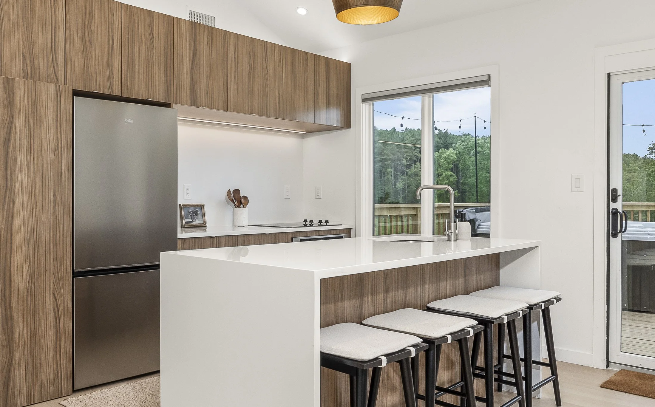 Modern kitchen with white island and wooden cabinets, four bar stools at the breakfast bar, stainless steel refrigerator, large window and sliding door showing outdoor deck and greenery.