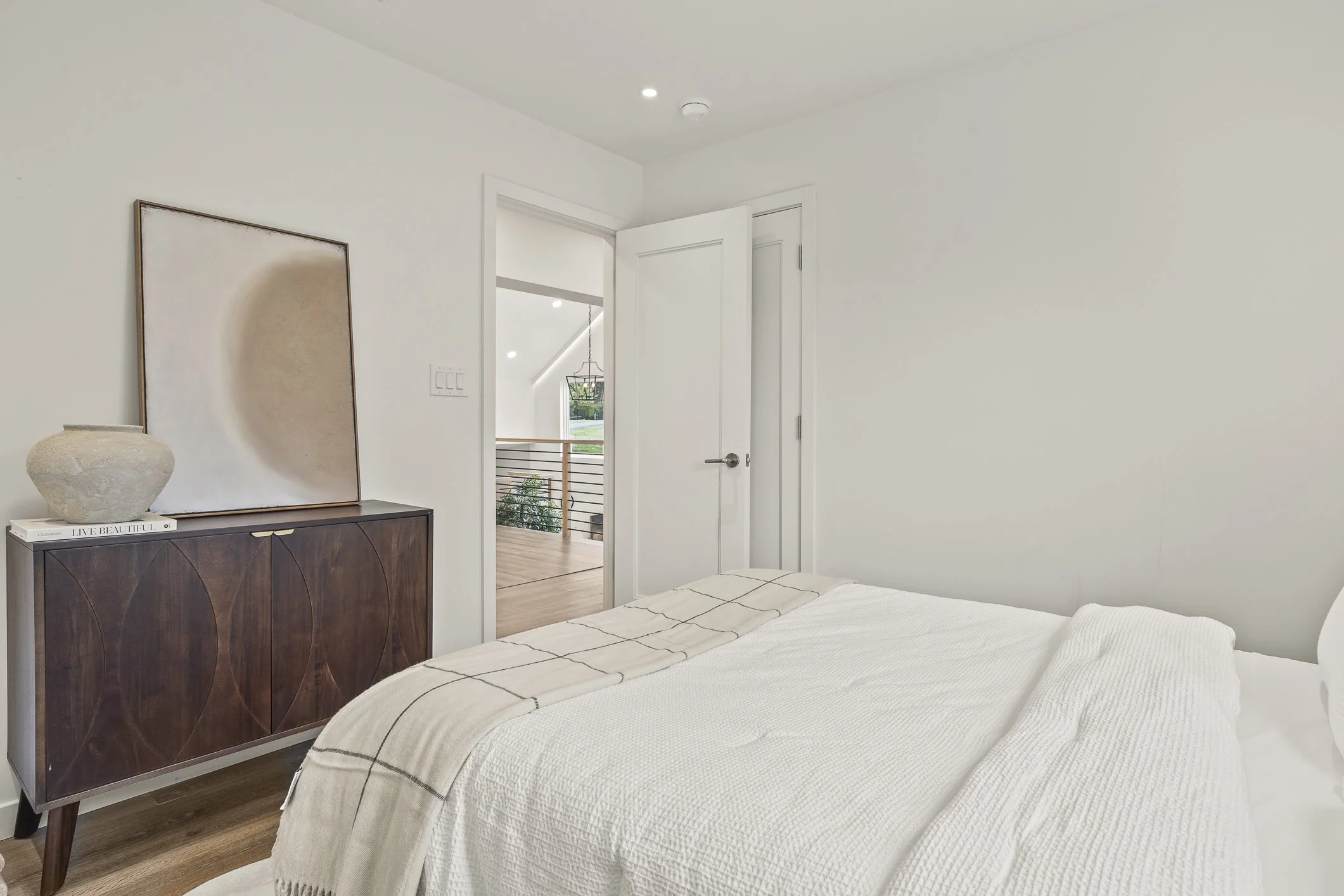A bedroom with white walls, a bed with white linens, a wooden sideboard with a large abstract art piece and a decorative vase, open door leading to an upstairs hallway with a wooden railing and a hanging light fixture, and hardwood floors.