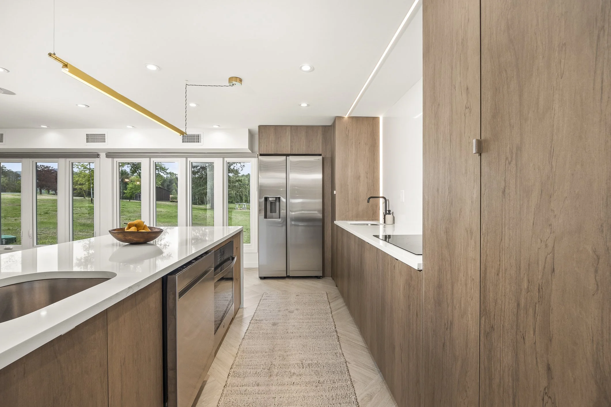 Modern kitchen with wooden cabinets, stainless steel refrigerator, white countertops, and a view of a green outdoor yard through large windows.