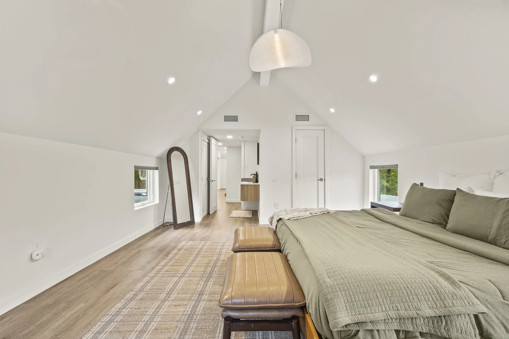 A bedroom with a sloped ceiling, white walls, hardwood floors, and a bed with green bedding. There are small windows on each side, a standing mirror, and a lamp fixture hanging from the ceiling.