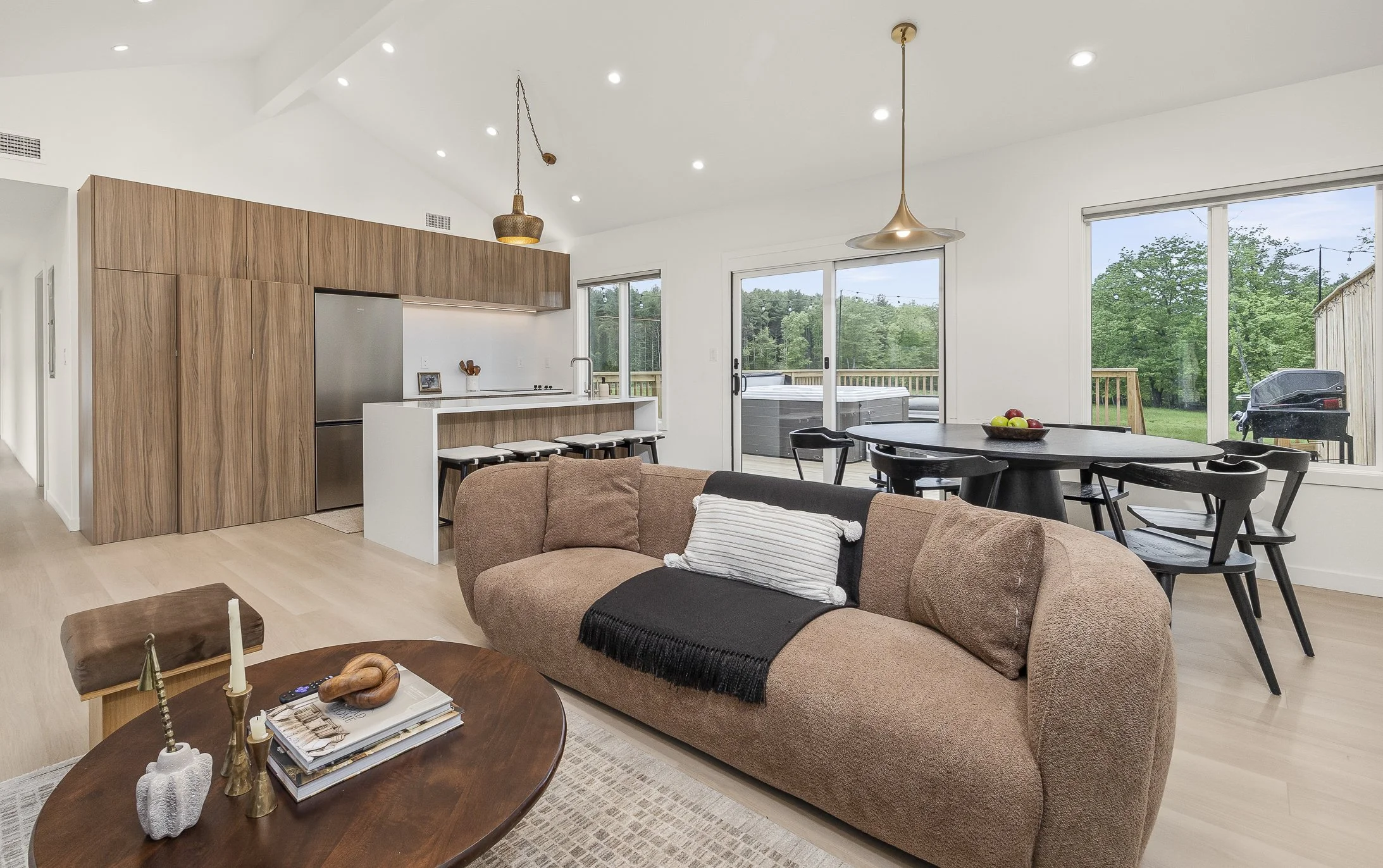 Open-concept living room and kitchen with minimalistic decor, wooden cabinets, black dining table with chairs, sliding glass door leading to a deck, and large windows overlooking a green backyard.