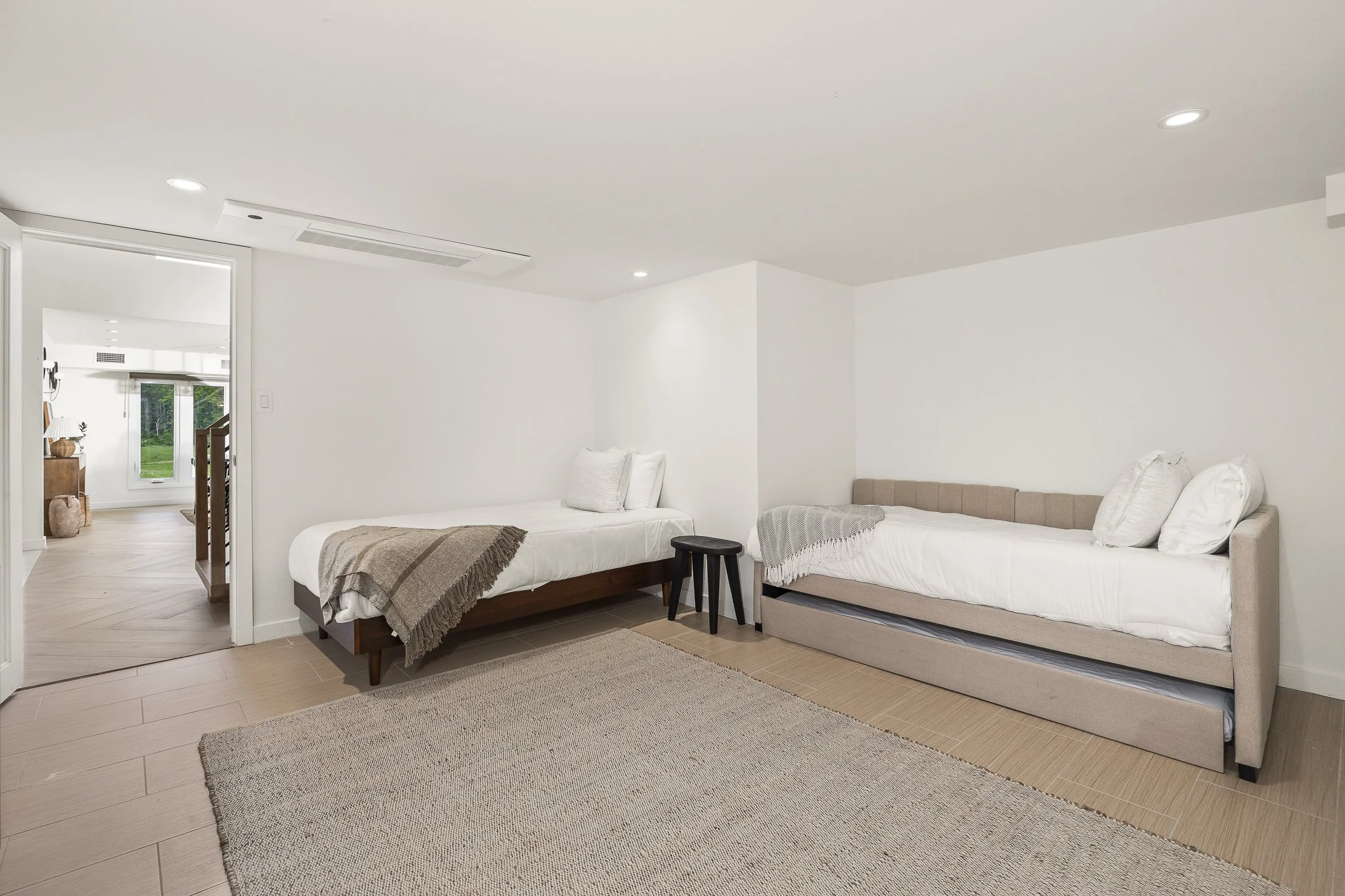 A minimalistic bedroom with two single beds, one with a wood frame and the other with a beige upholstered frame, a black stool, a textured beige rug, and white walls, with a view into another bright room with a sliding glass door.