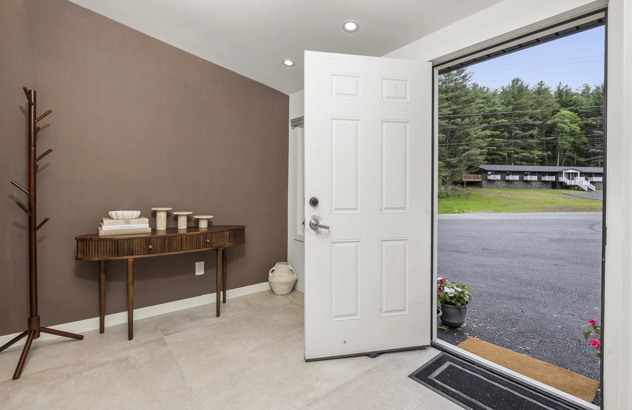 Entryway with open front door, view of outdoor driveway and house with trees in the background, indoor side table with decorative items, potted plants outside, and a brown wall with a coat rack.