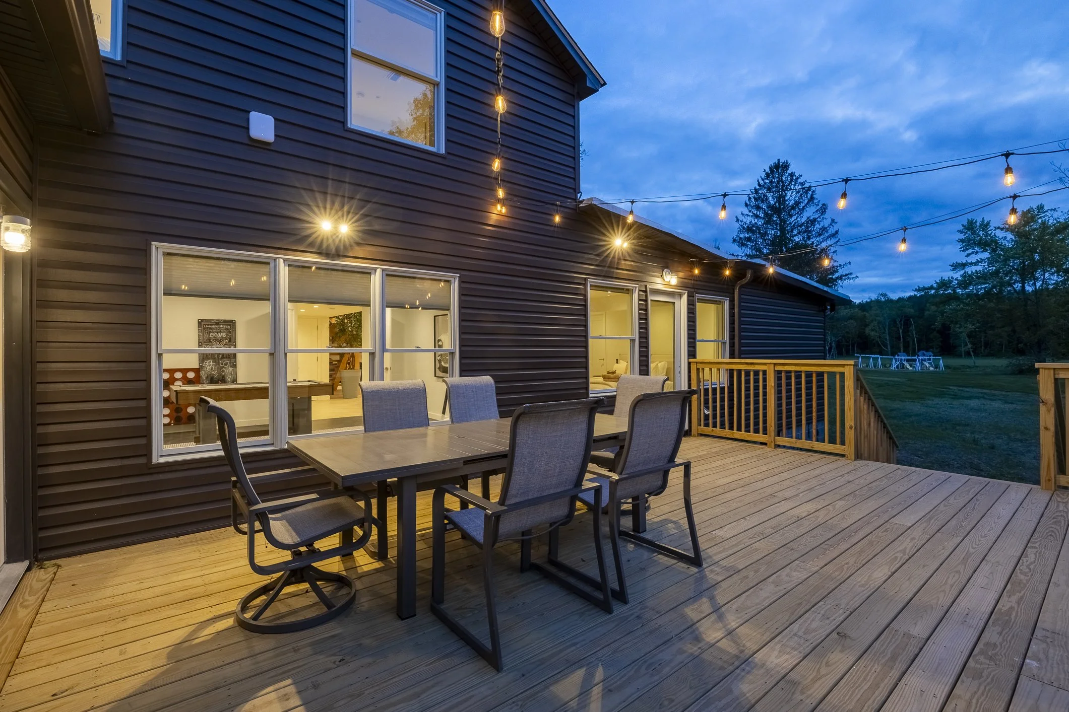 A wooden outdoor deck with a dining table and six chairs outside a house, decorated with string lights at dusk, with a grassy backyard and trees in the background.