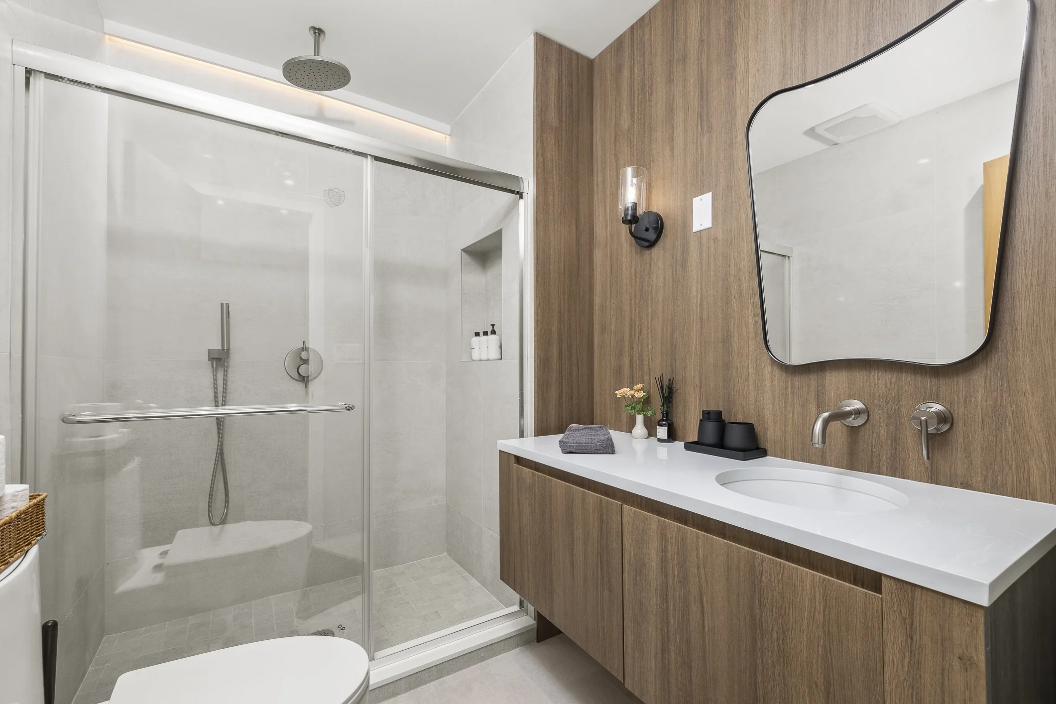 Modern bathroom with a glass-enclosed shower, wooden vanity, white countertop, and a uniquely shaped mirror.