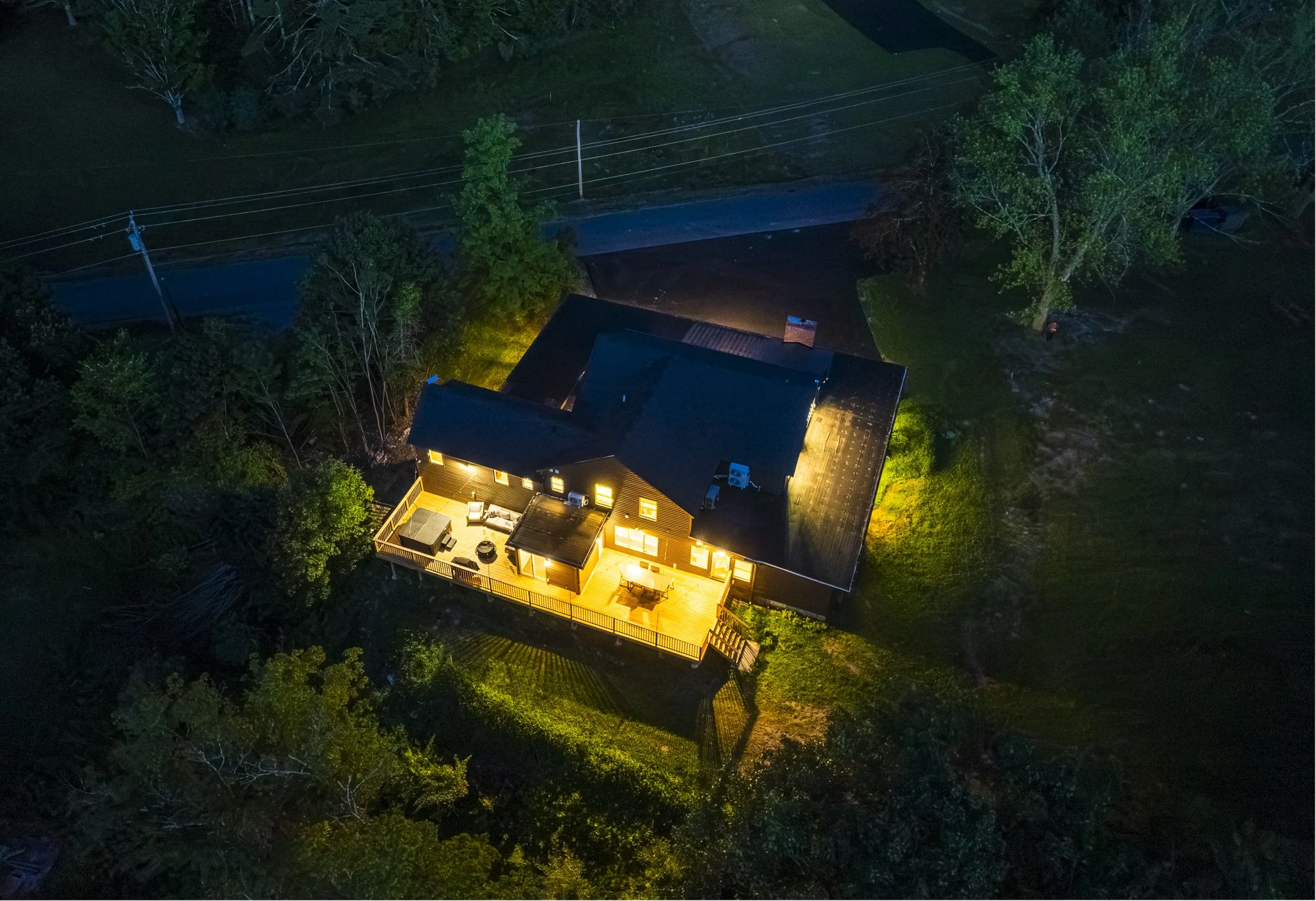 A house at night with outdoor lighting on a deck, surrounded by trees and grass, and a road in the background.
